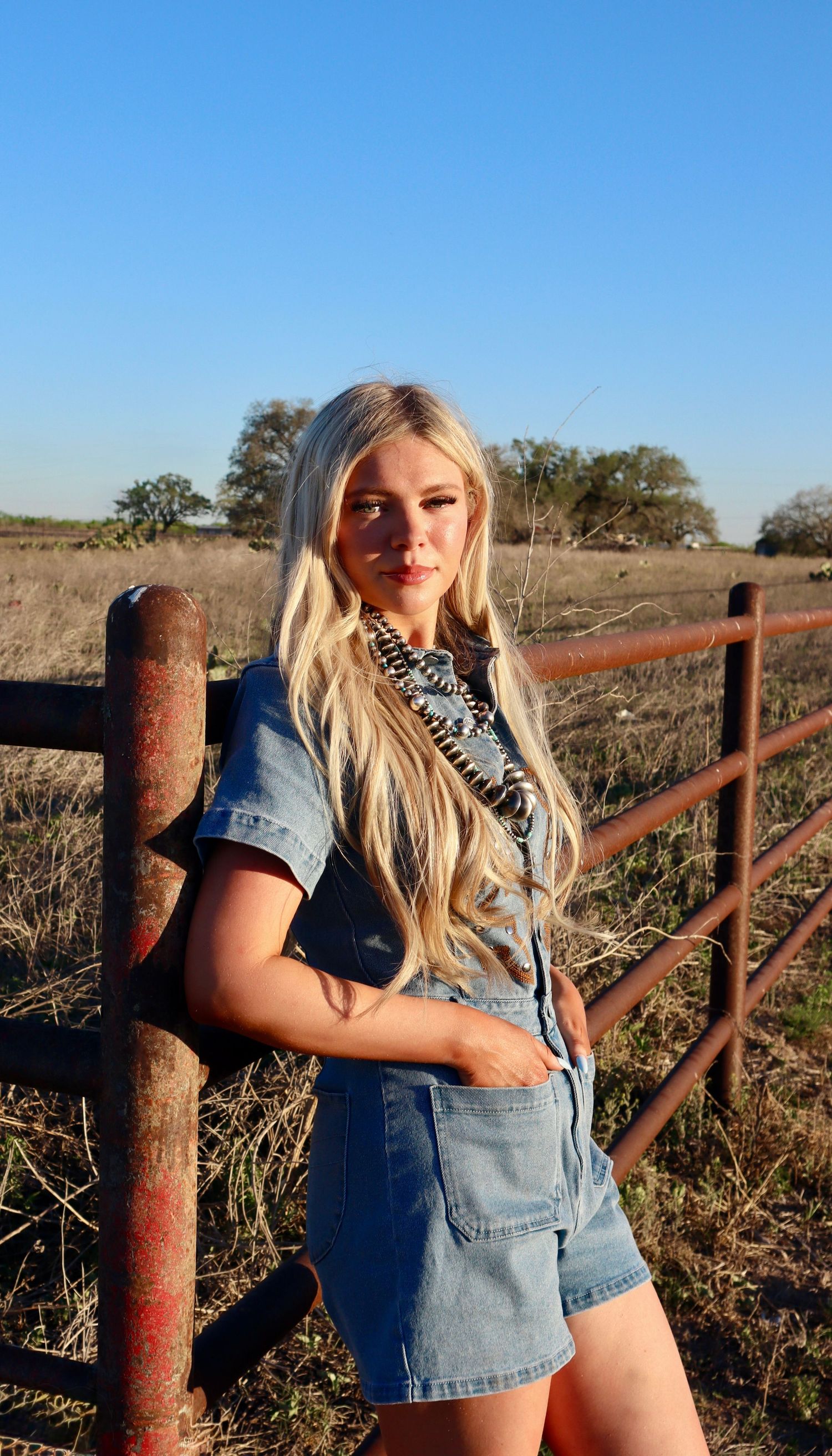 Person in denim overalls poses against rustic wooden fence in rural field at sunset.