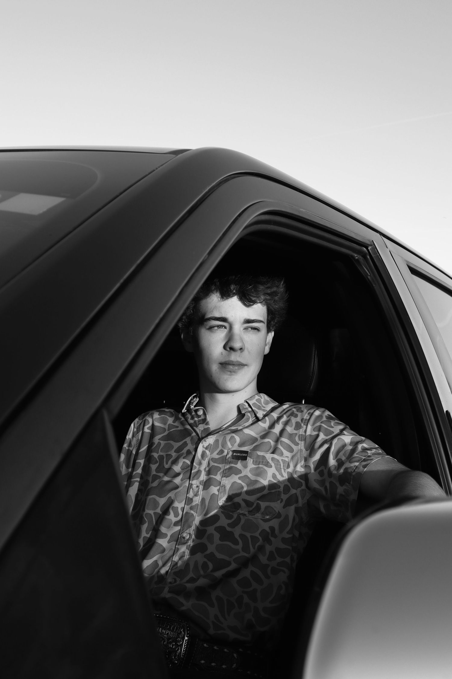 Black and white portrait of someone sitting in a car wearing a patterned button-up shirt.