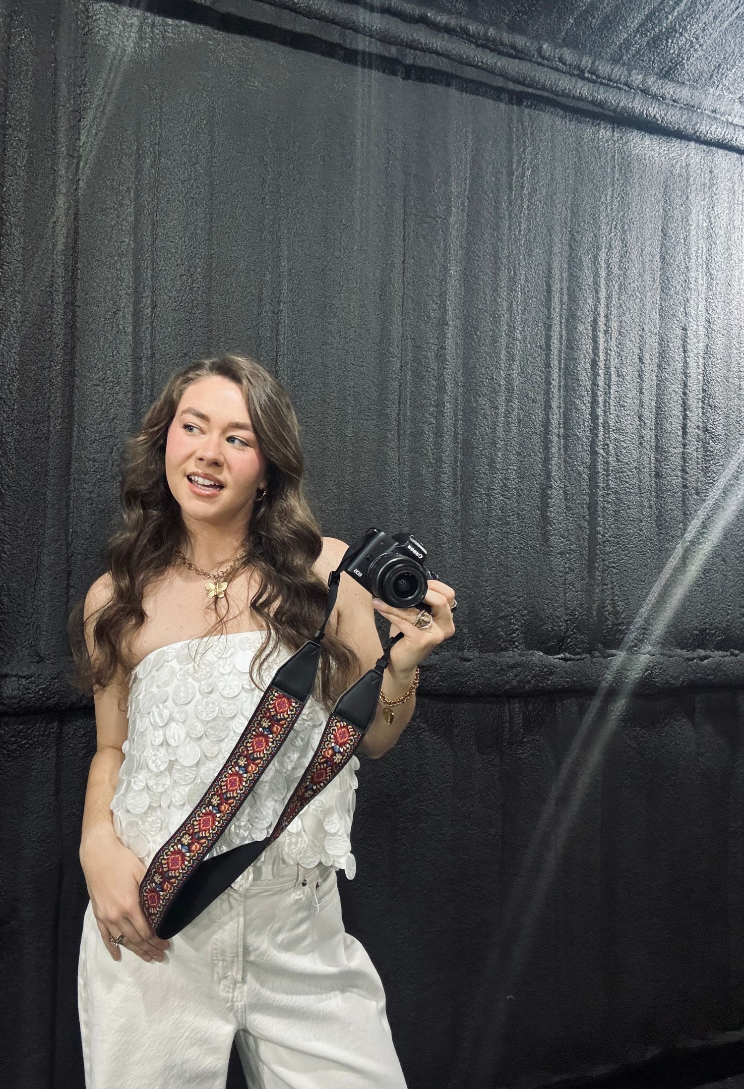 A person in a white outfit poses with a camera and embroidered strap against a dark backdrop.