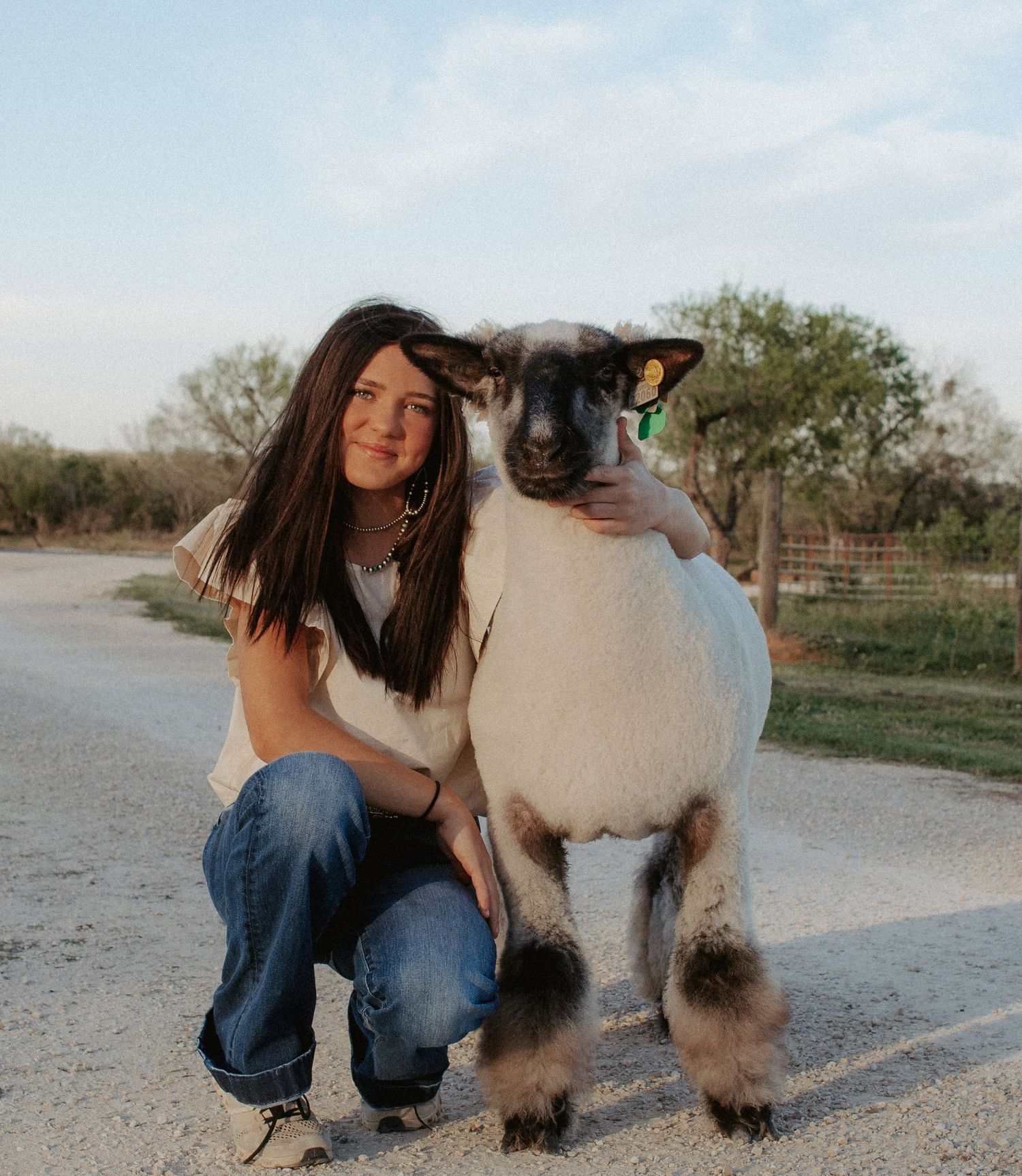 A person in blue jeans kneels beside a fluffy white and black sheep on a dirt path in a rural setting.
