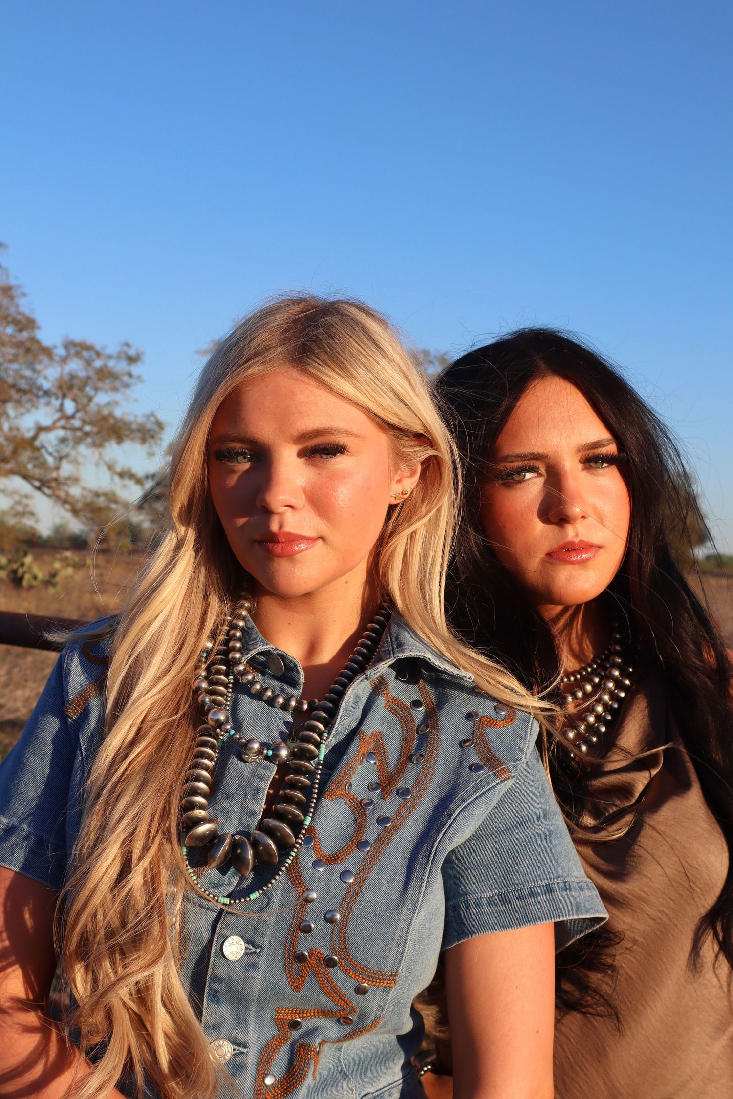 Two women in western style outfits with turquoise necklaces pose outdoors at sunset against a blue sky.