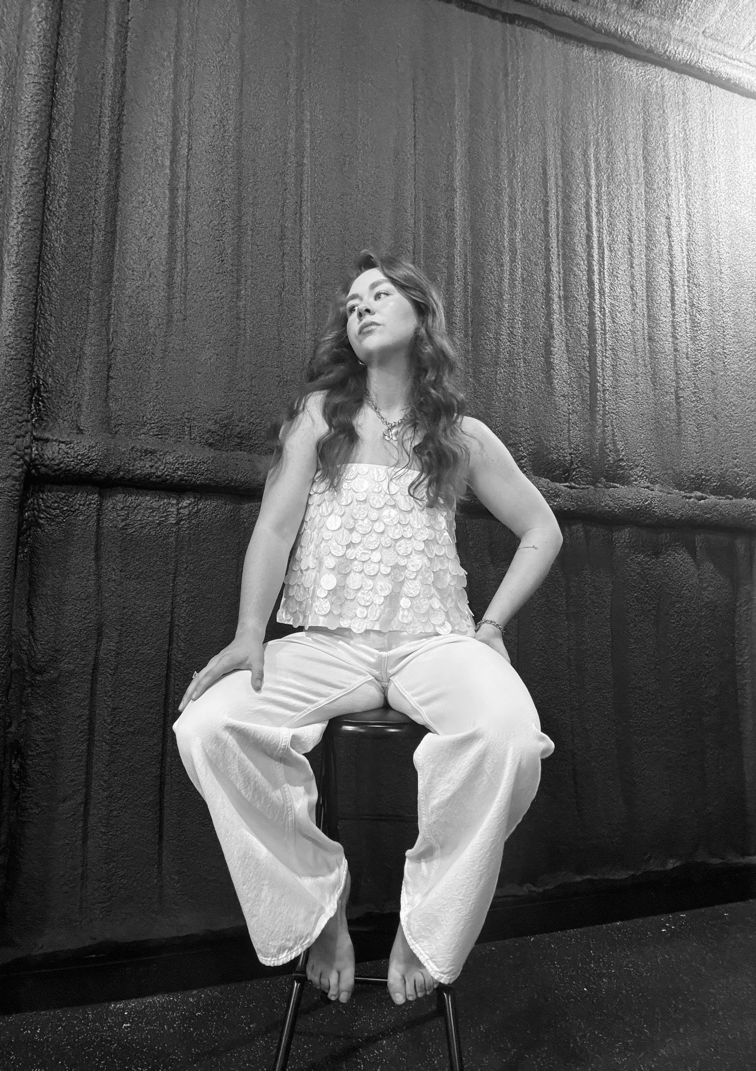 A person in a white dress sits on a chair in a moody black and white photograph with dramatic lighting.