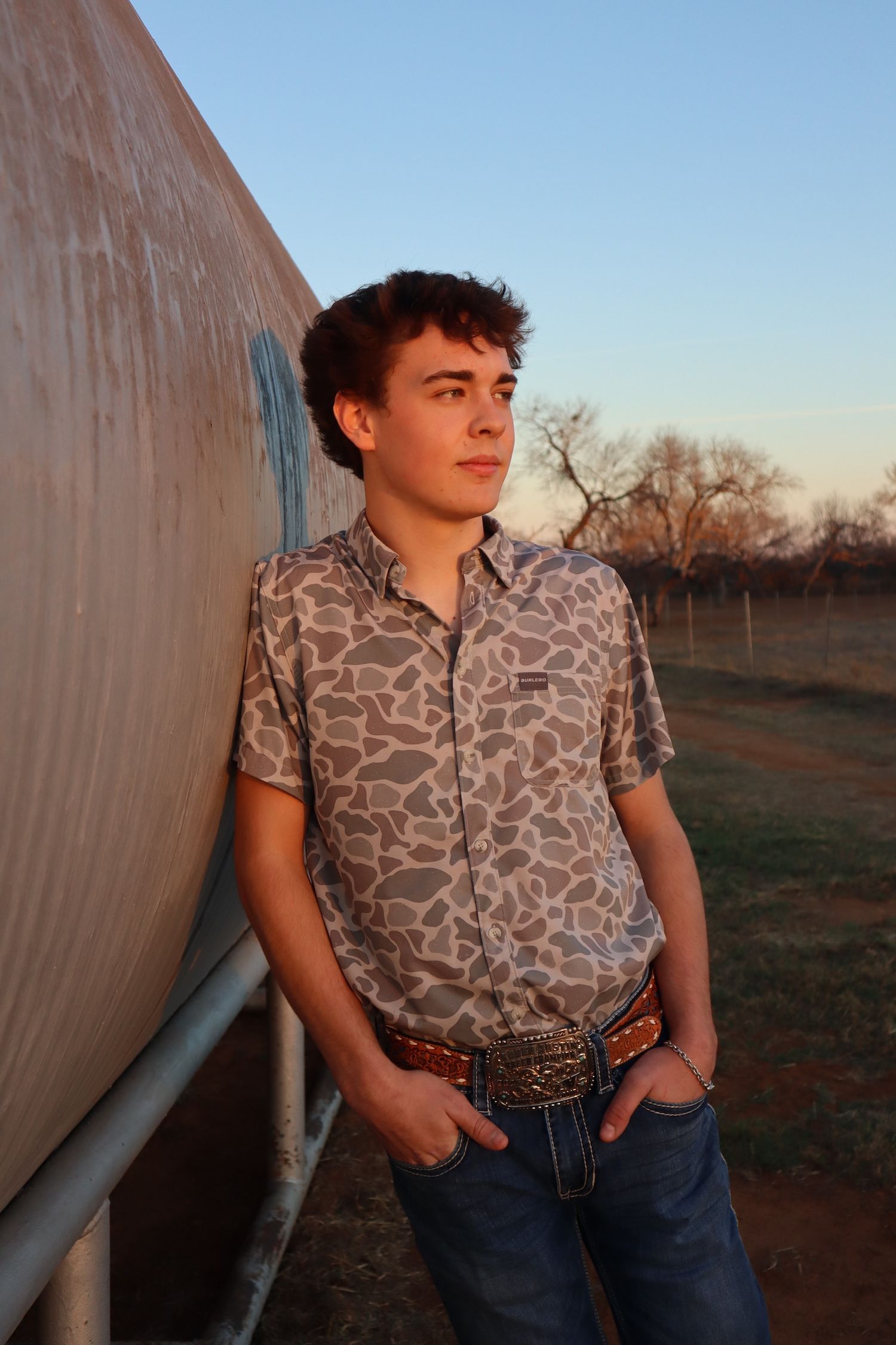 Person in patterned button-down shirt and jeans leans against wall during sunset in rural setting.