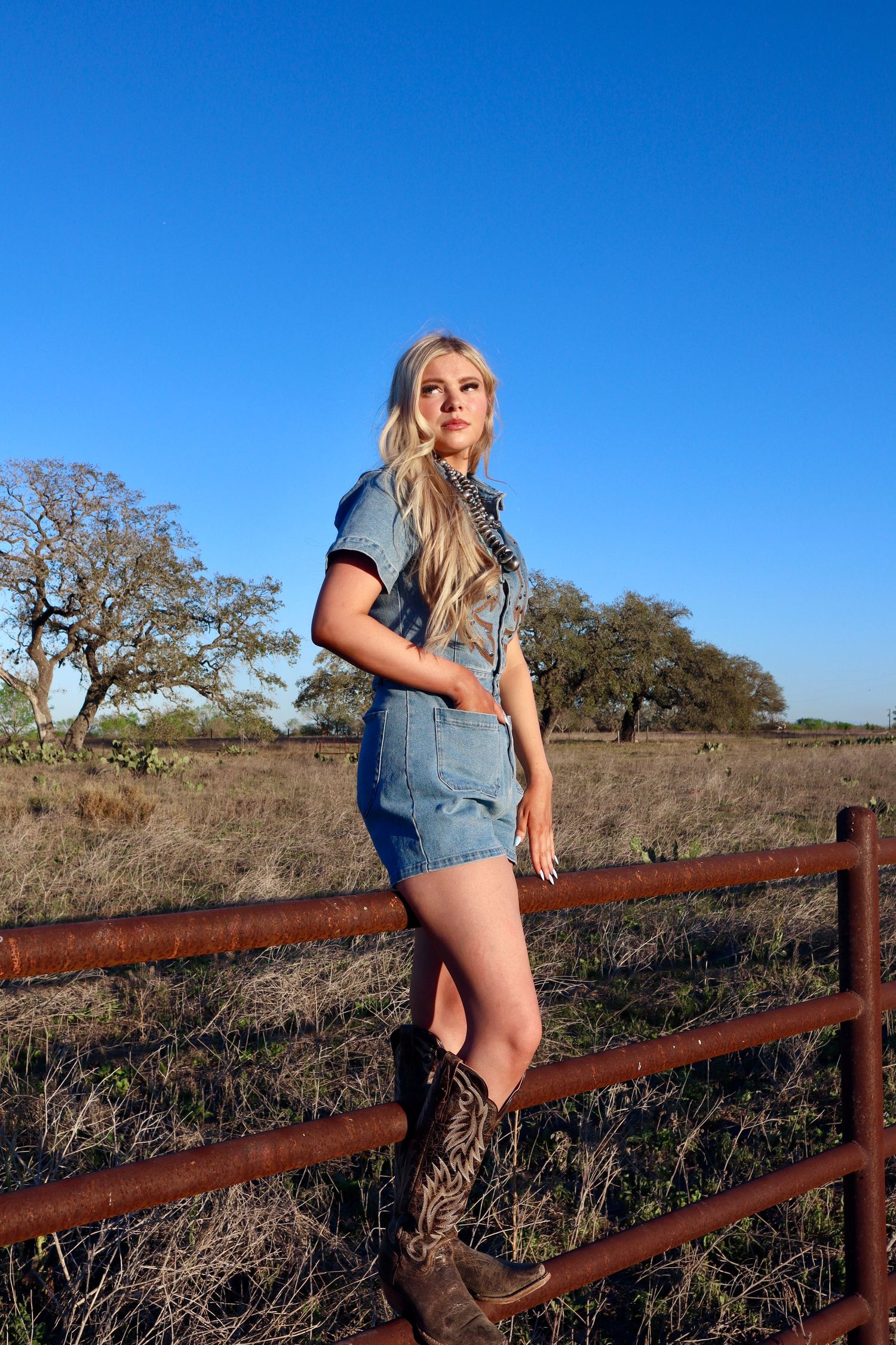Person in denim and boots sits on rustic wooden fence against bright blue sky and rural landscape.