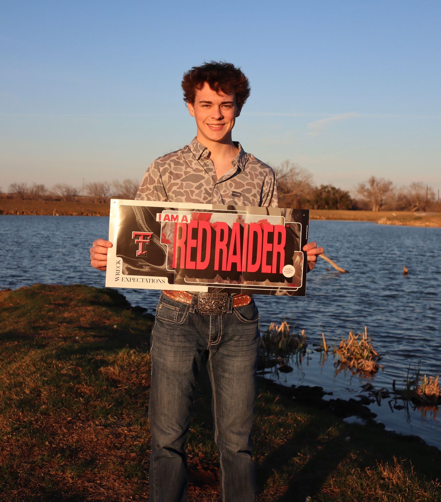 Someone holds up a vintage-style 'Red Raiders' sign while standing by a lake during sunset.
