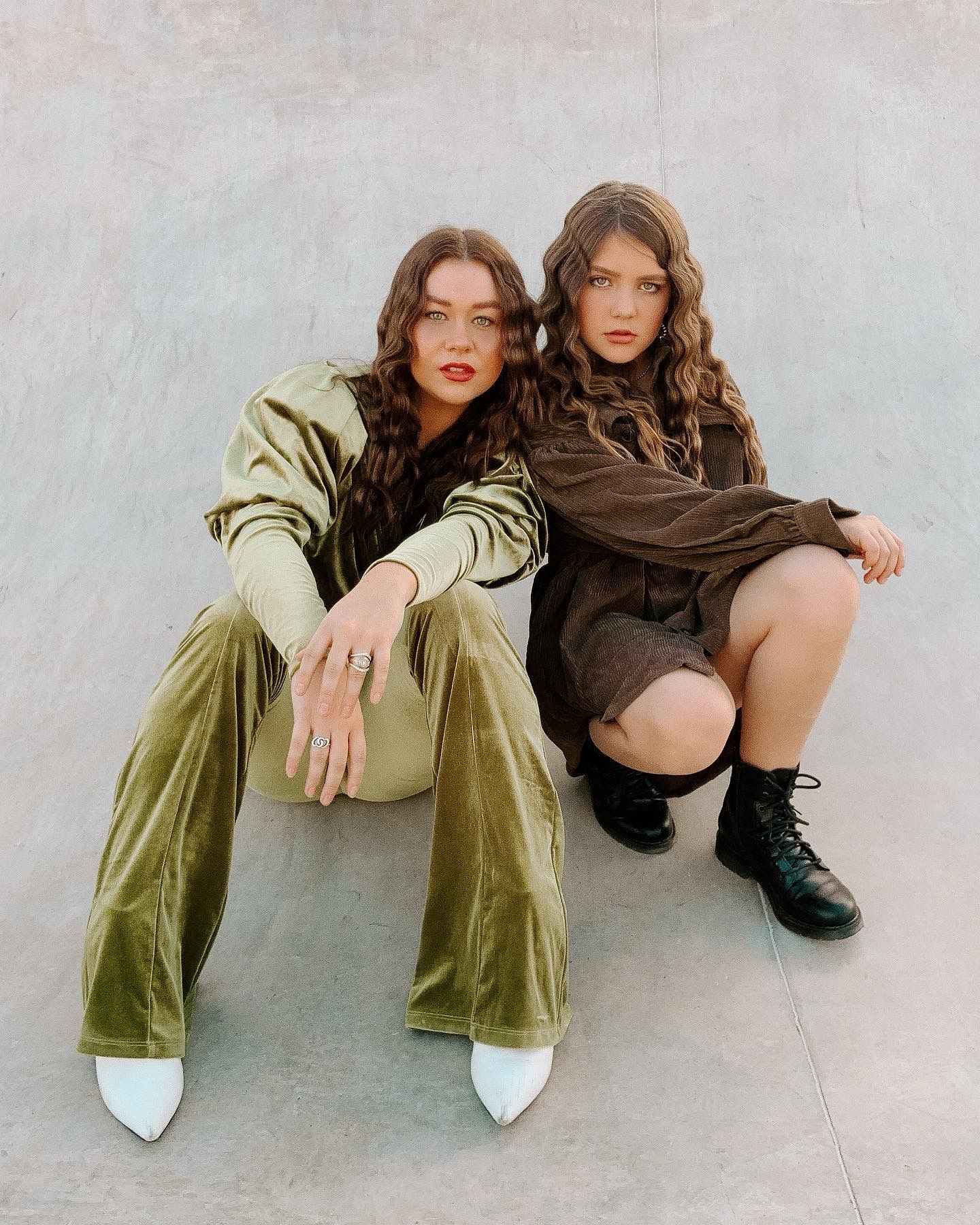 Two people in contrasting outfits of olive green velvet and dark brown pose casually on a light gray floor.