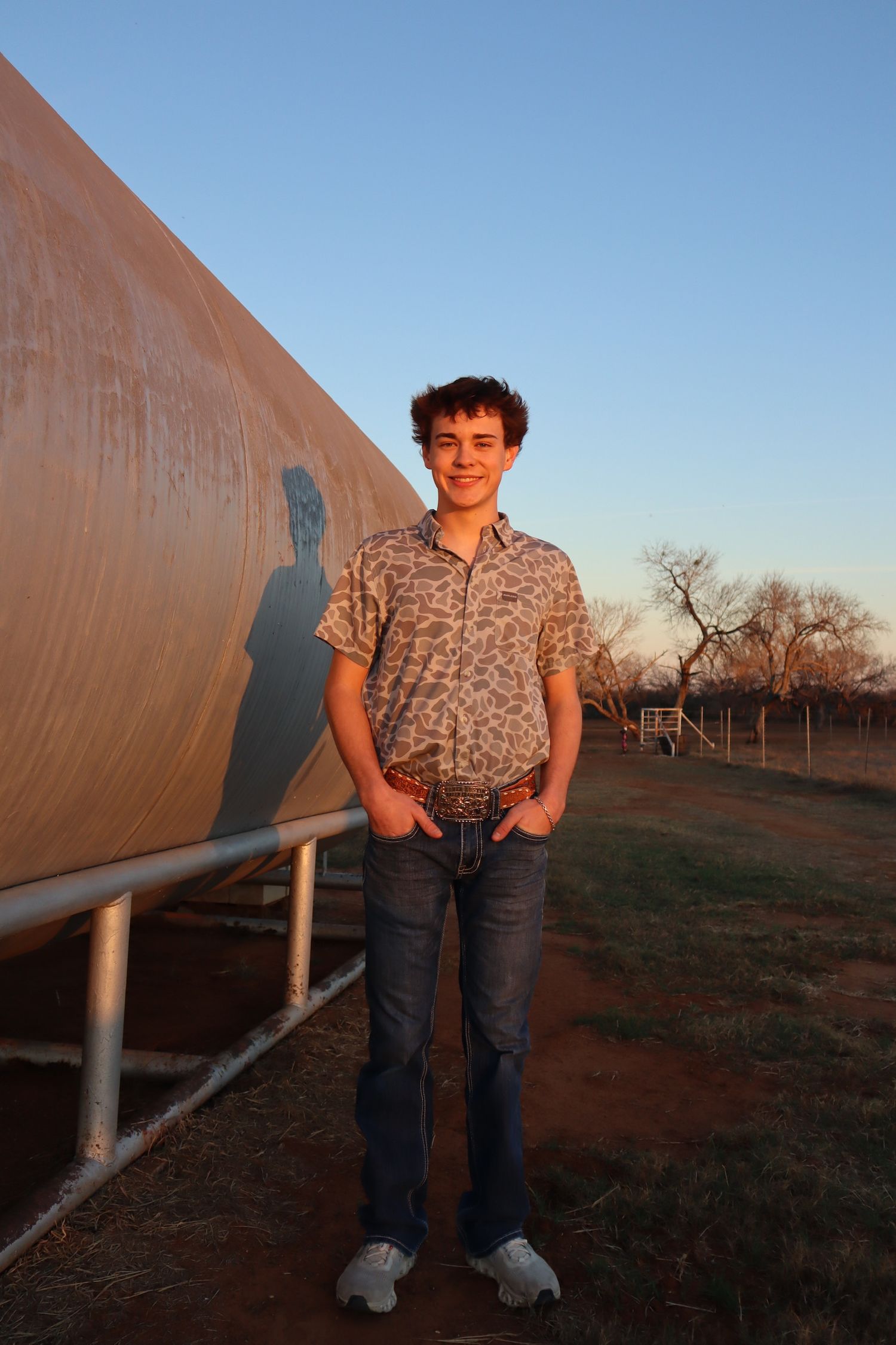 A person stands against a metal fence structure in an open field at sunset.