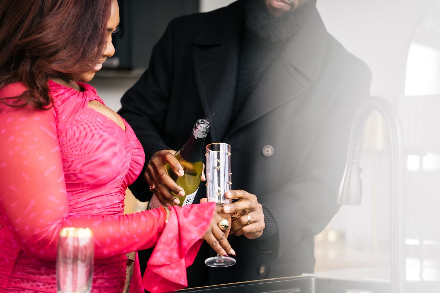 Couple pouring champagne together in kitchen, woman in pink dress.