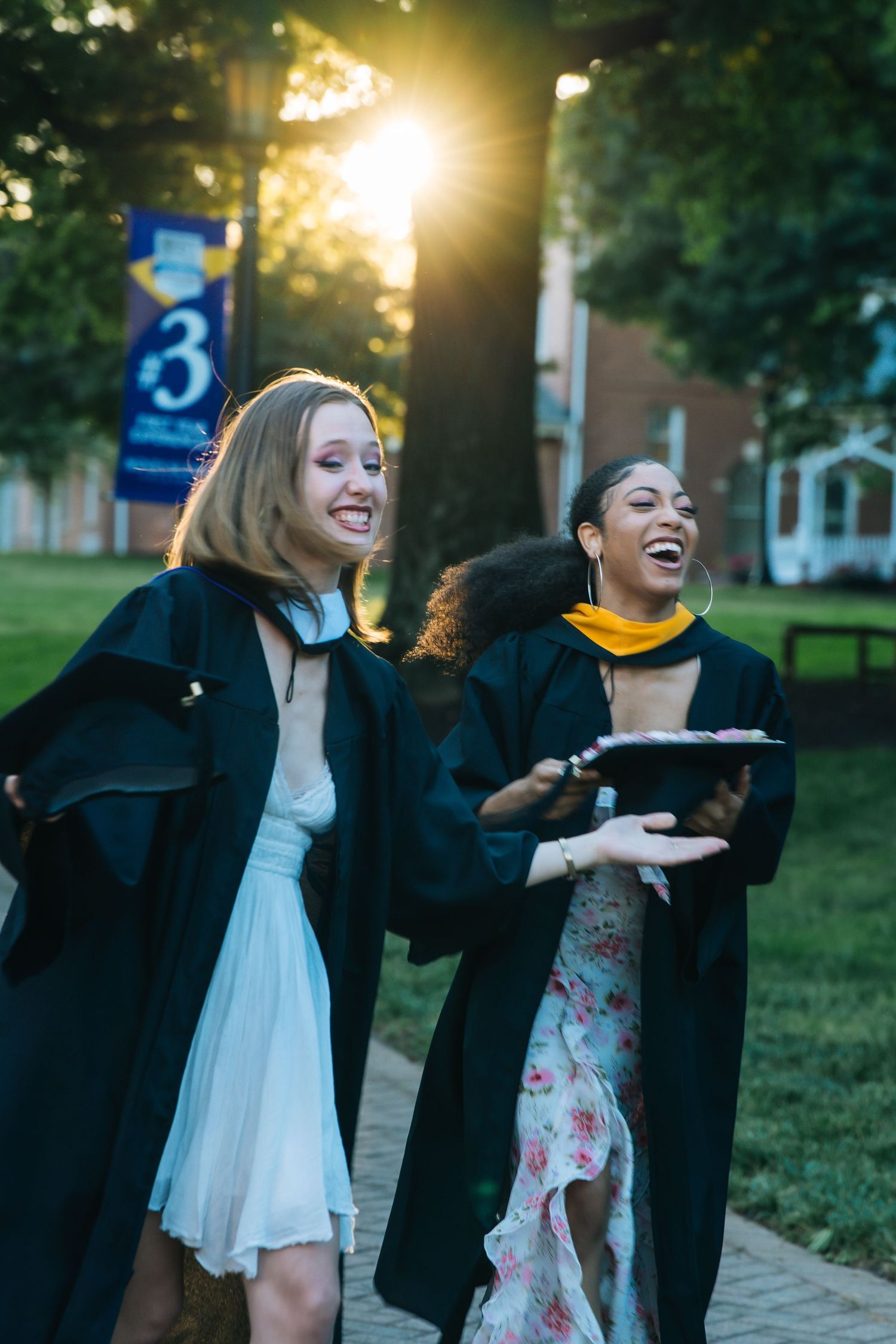 Two laughing female graduates in black robes celebrate outdoors in sunlight.