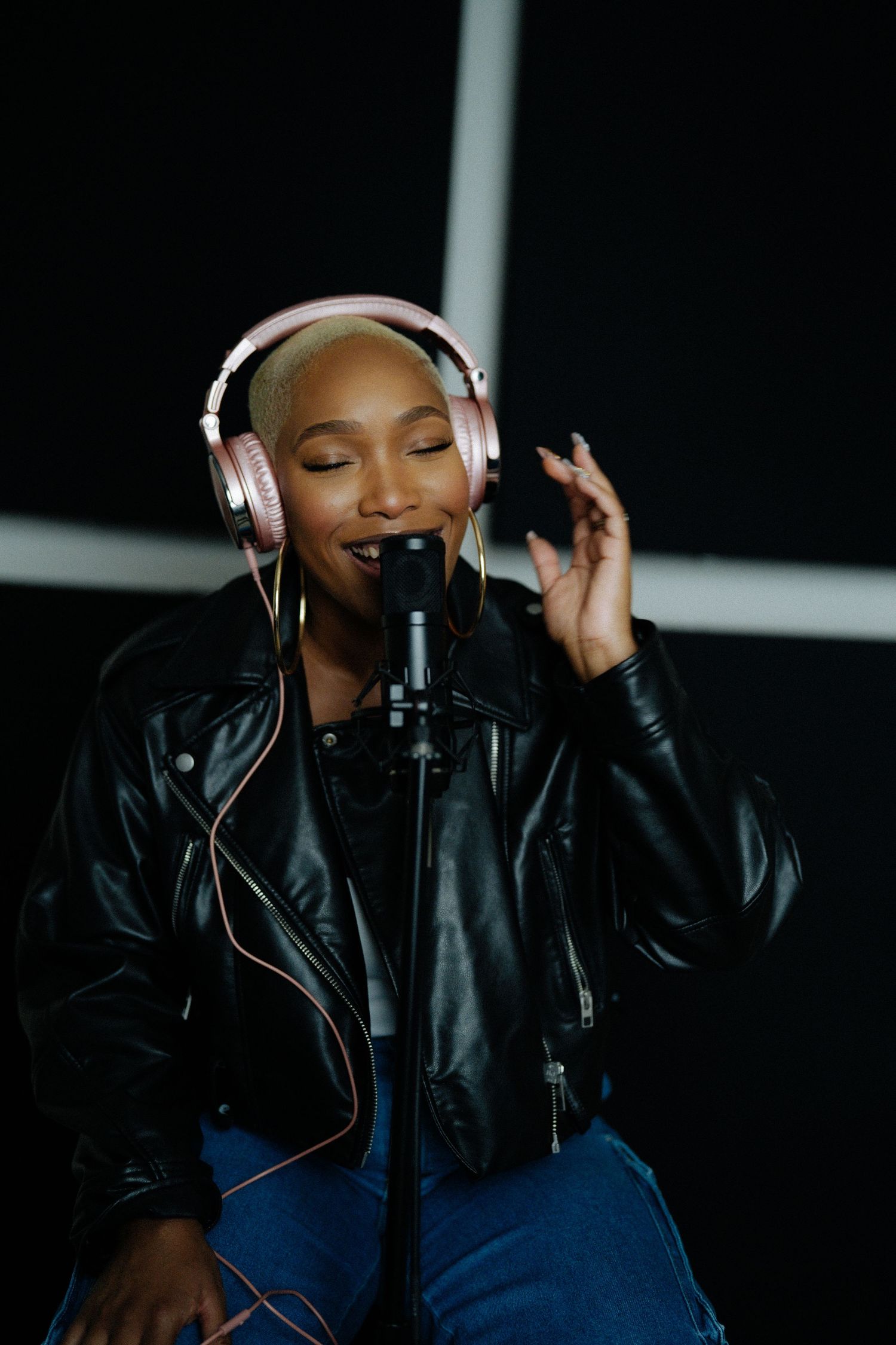 Female vocalist with shaved head singing in recording studio wearing pink headphones and black leather jacket.