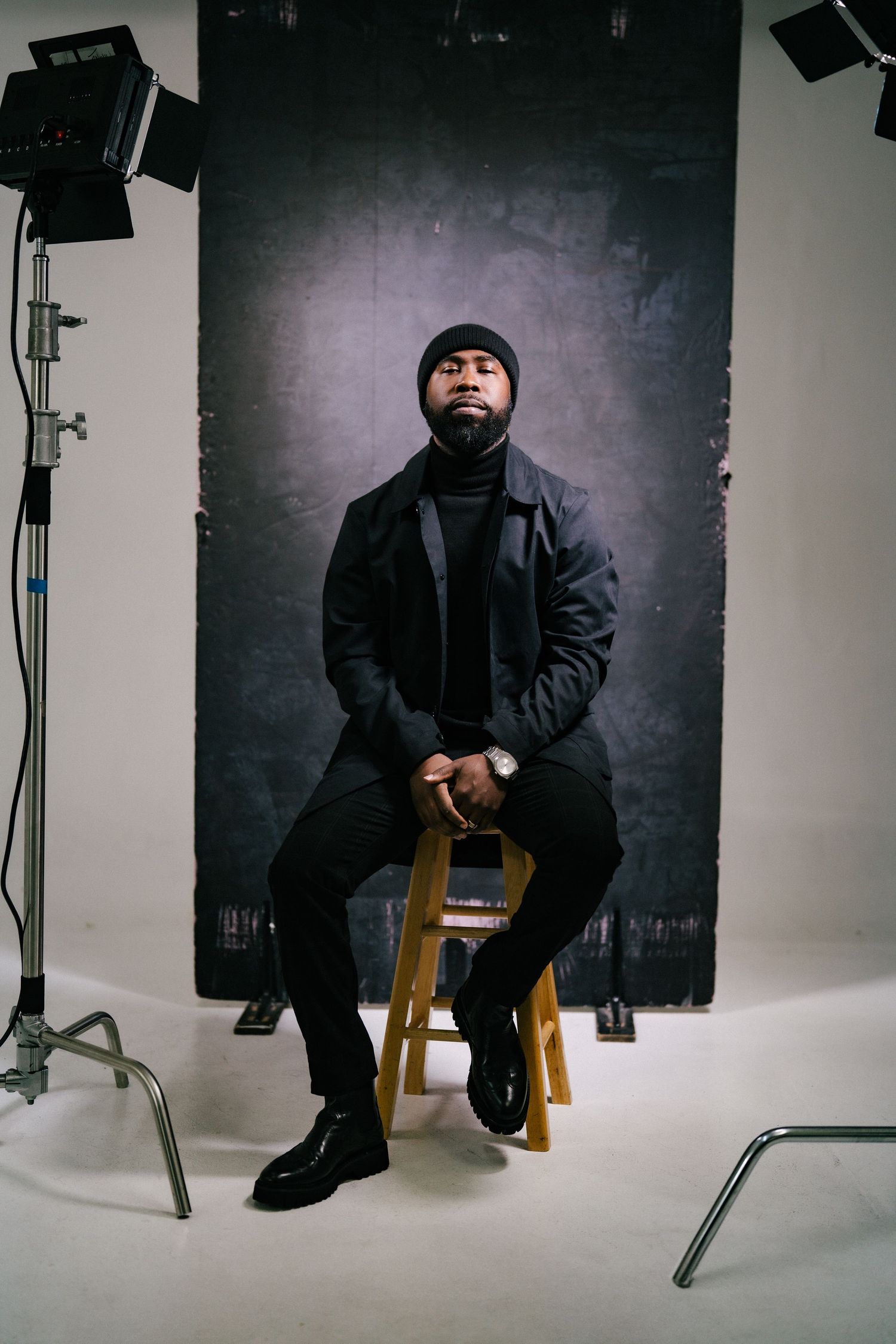 Black man in all-black outfit sitting on a stool in a photography studio with professional lighting.