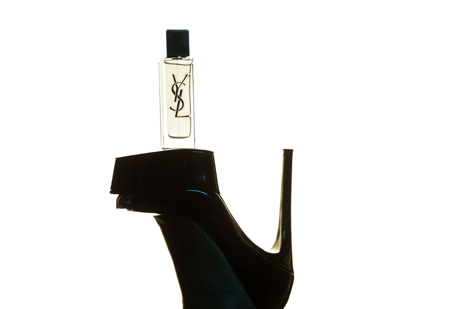 Small YSL perfume bottle with black cap balanced on the ruffled ankle cuff of a black high-heeled shoe against a white background.