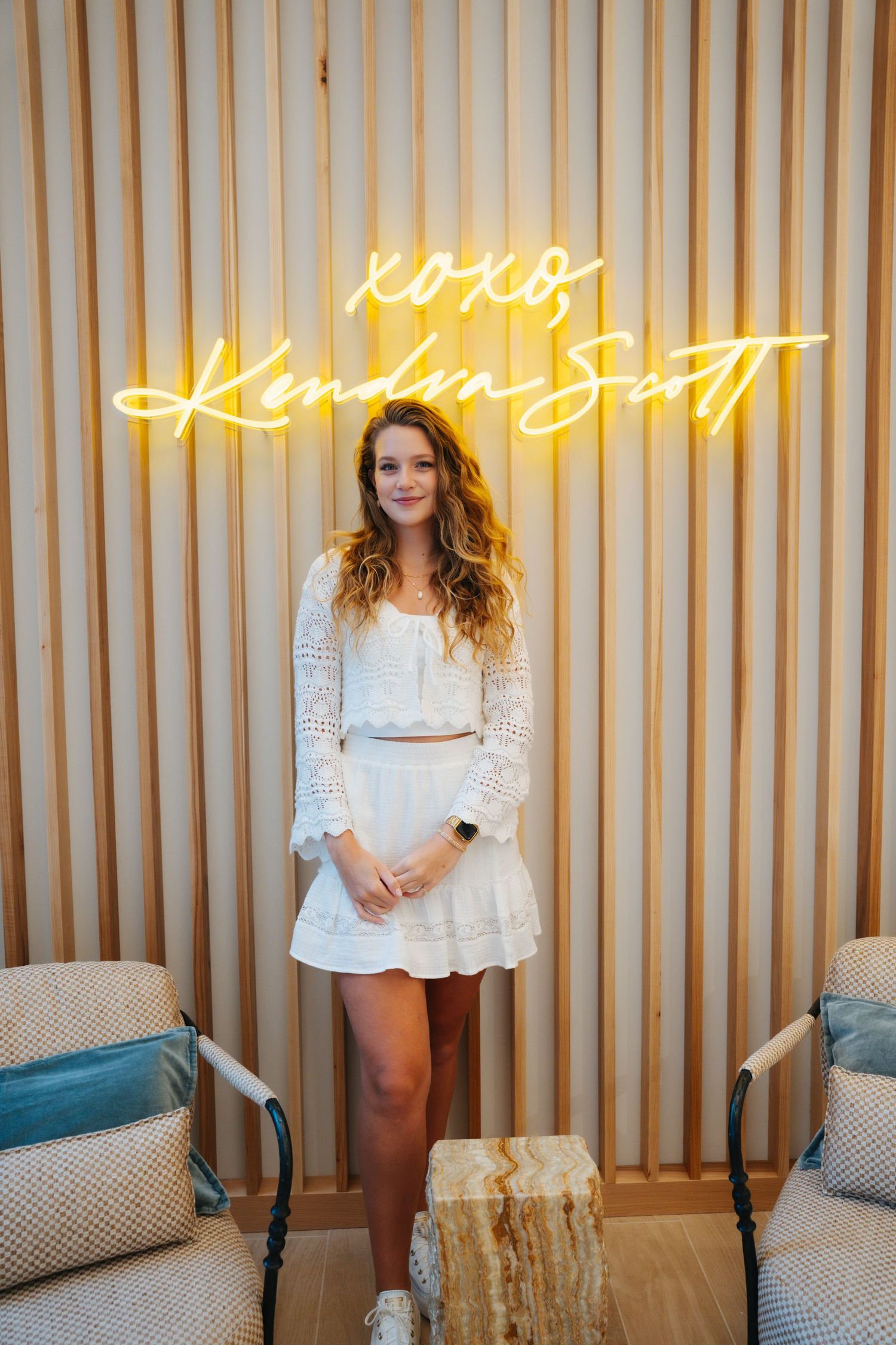 Woman in white lace co-ord set poses before a Kendra Scott neon sign on a wooden slat wall.