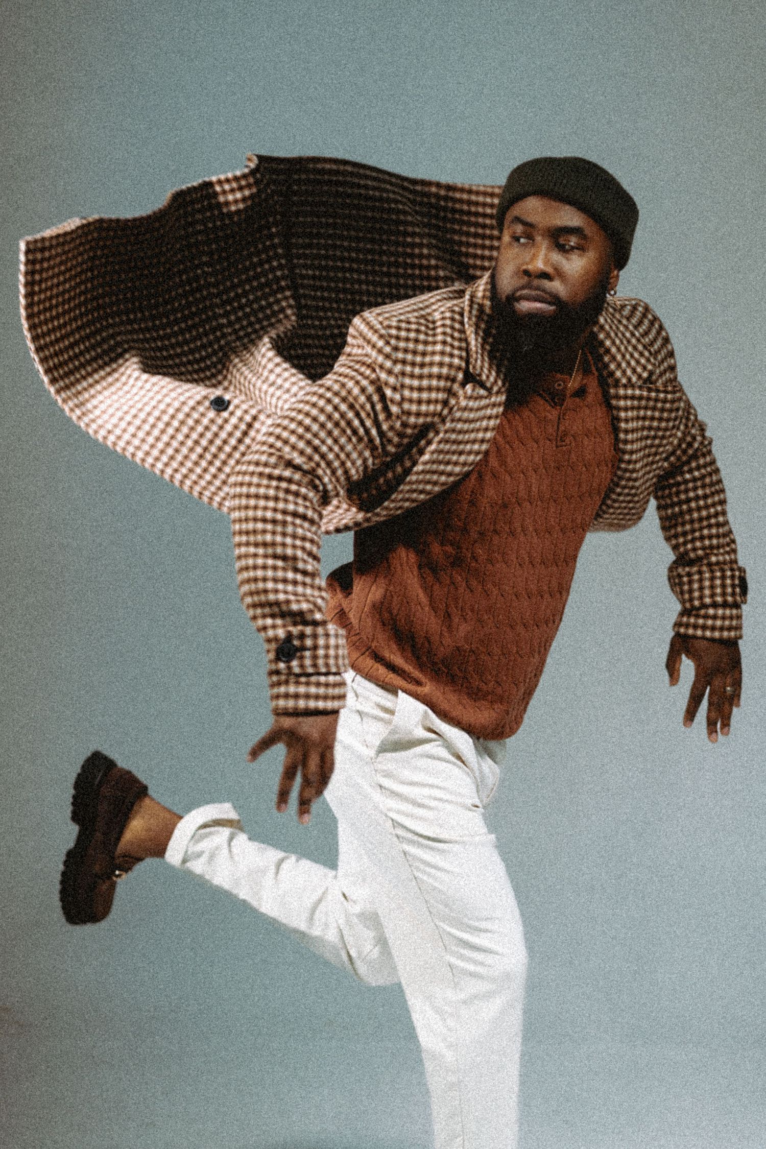Dynamic shot of a Black man in houndstooth coat and white pants jumping mid-air on a blue background.