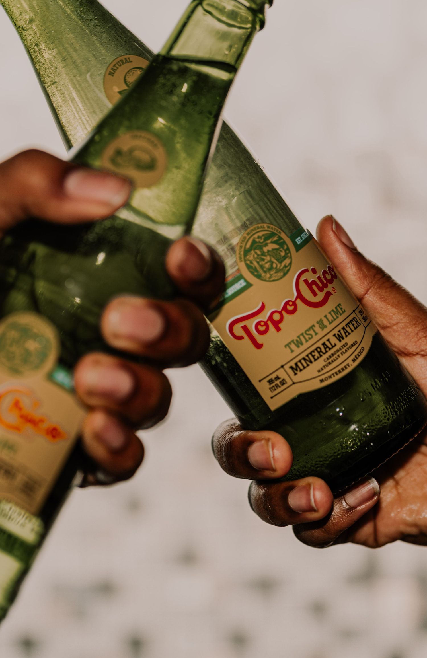 Two hands holding Topo Chico Twist of Lime mineral water glass bottles together.
