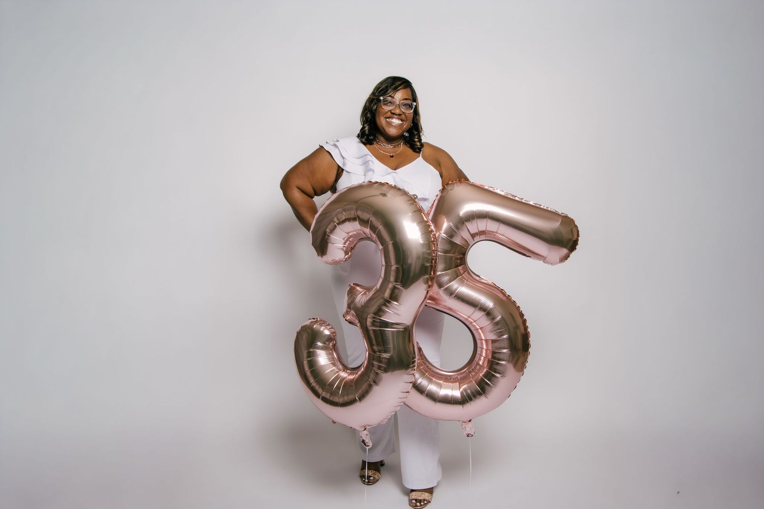 Woman celebrating 35th birthday holding rose gold number balloons in white outfit against white background.