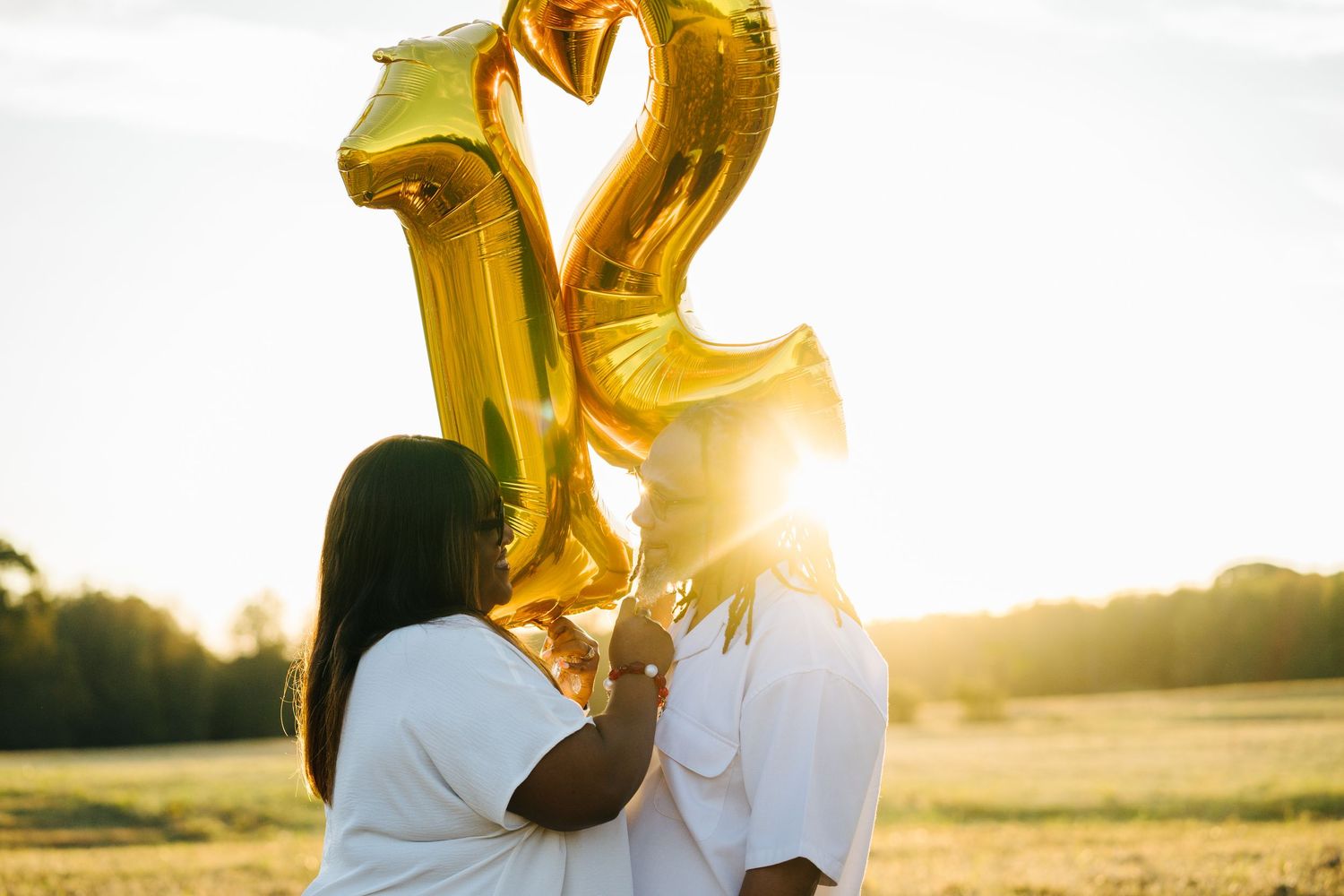Couple holding gold number balloons in a sunlit field during a golden hour engagement session.
