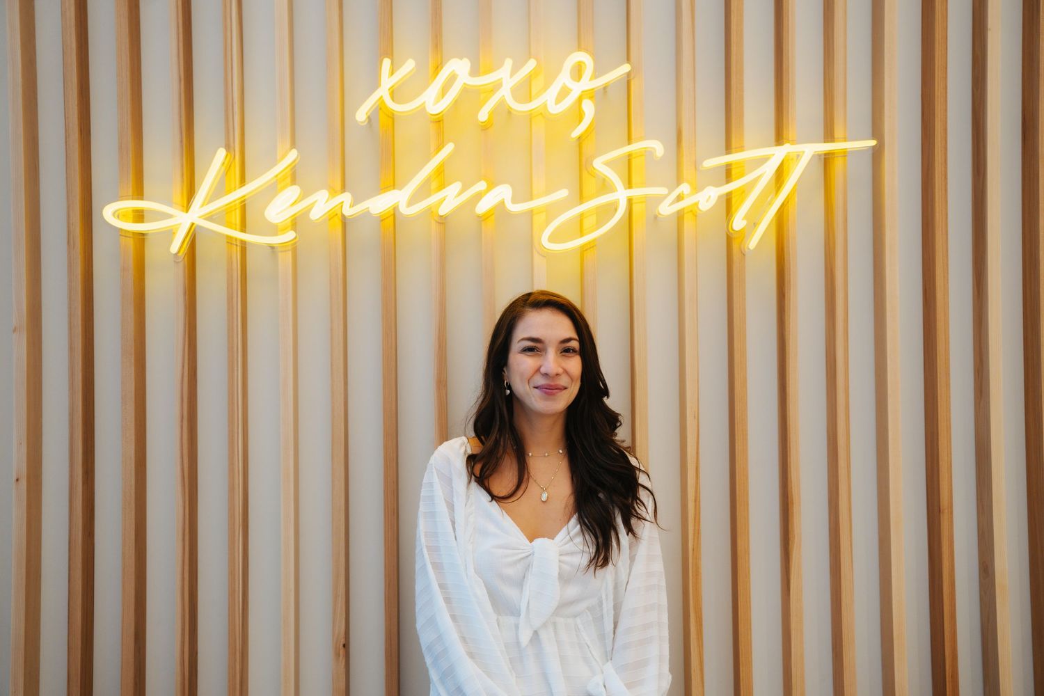 Dark-haired woman in white blouse smiling before XOXO Kendra Scott neon sign.