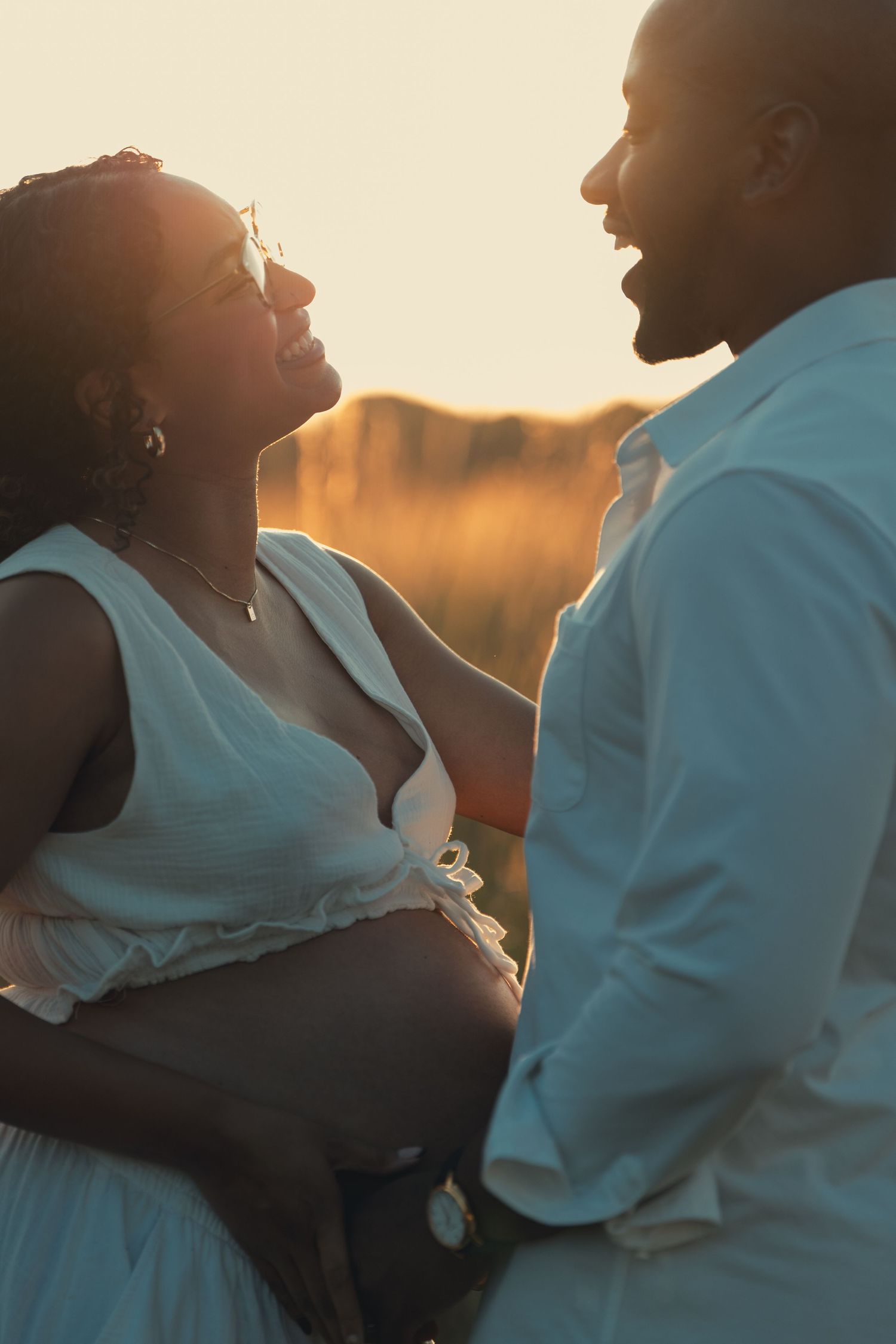 Couple laughs together during golden hour maternity photoshoot in open field.