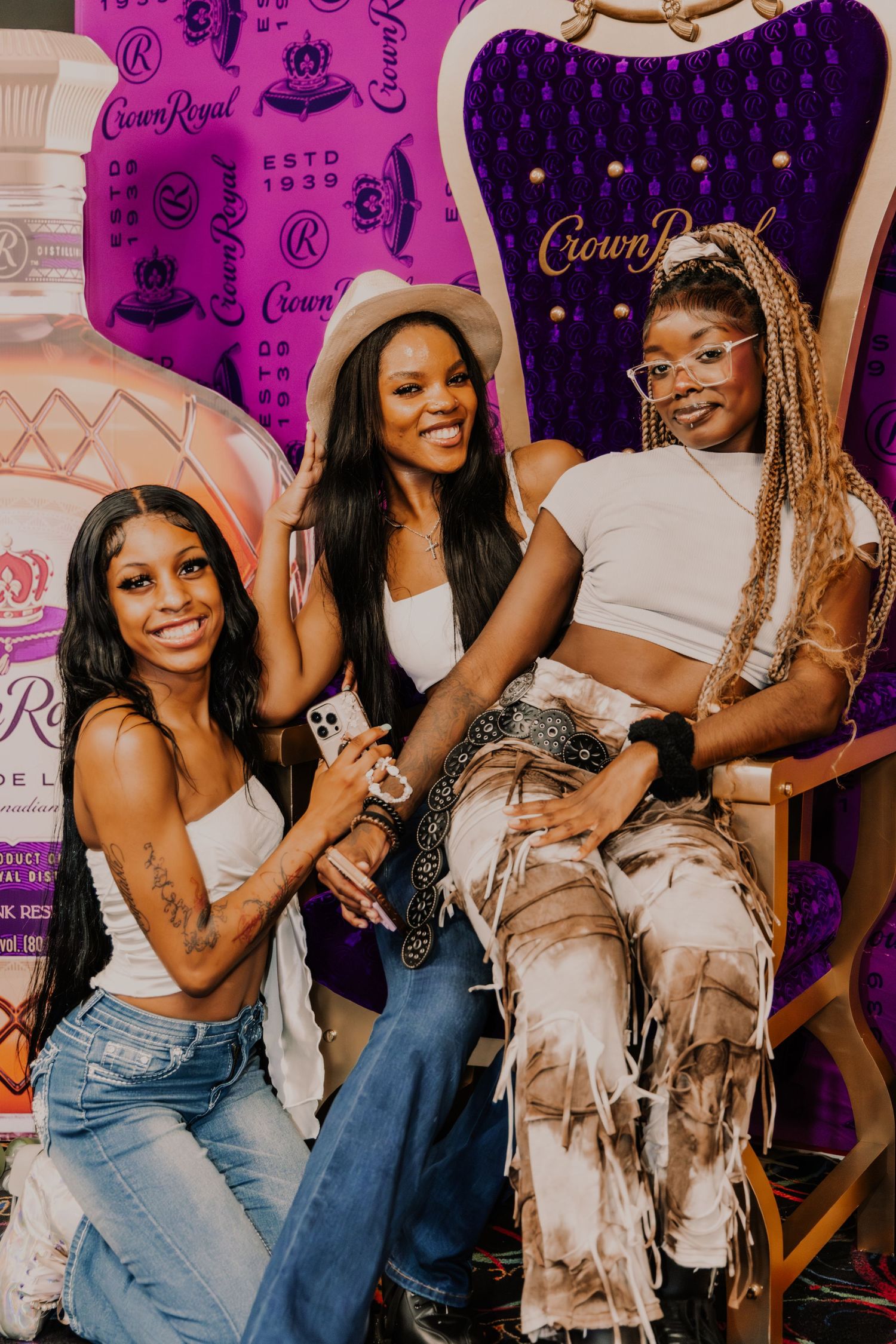 Three women pose and smile in front of a Crown Royal branded backdrop at a promotional event.