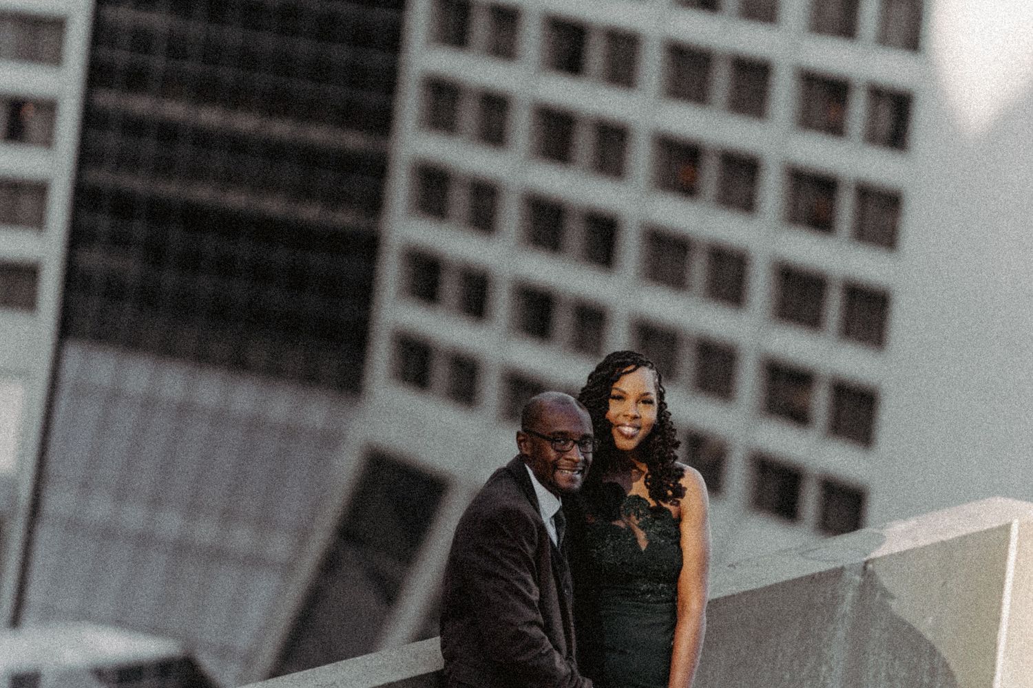 Couple in elegant formal attire posing together on urban rooftop with city buildings behind.