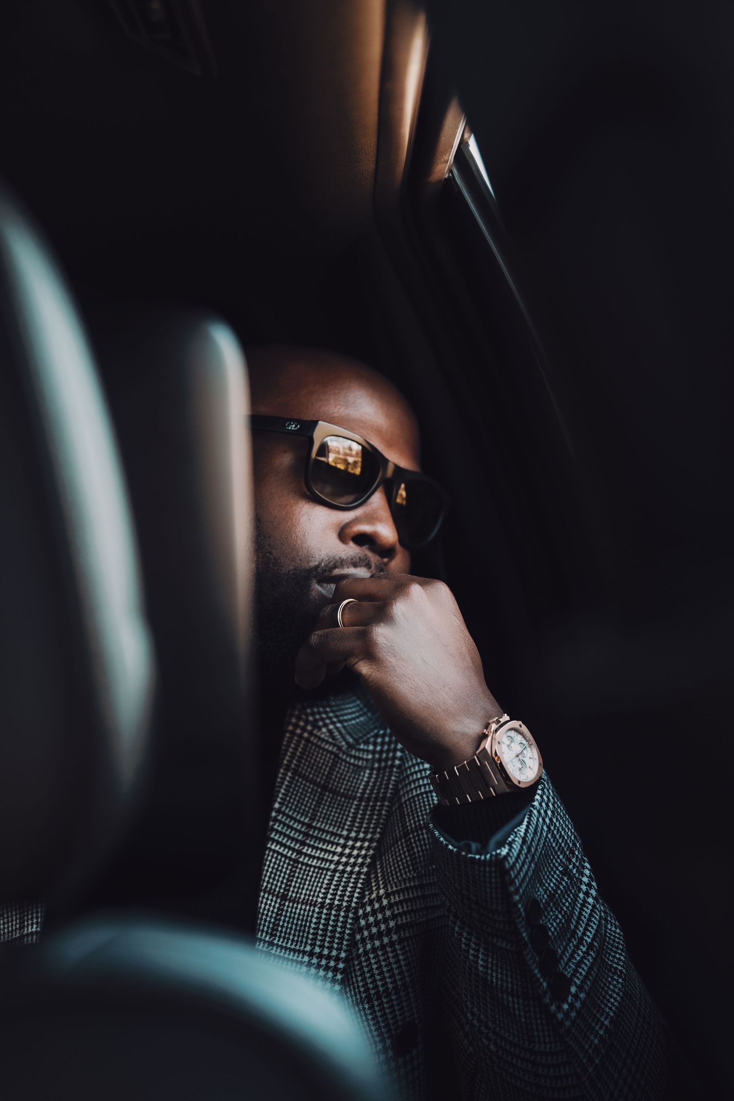 A stylish man in a plaid blazer and sunglasses sitting inside a luxury vehicle.