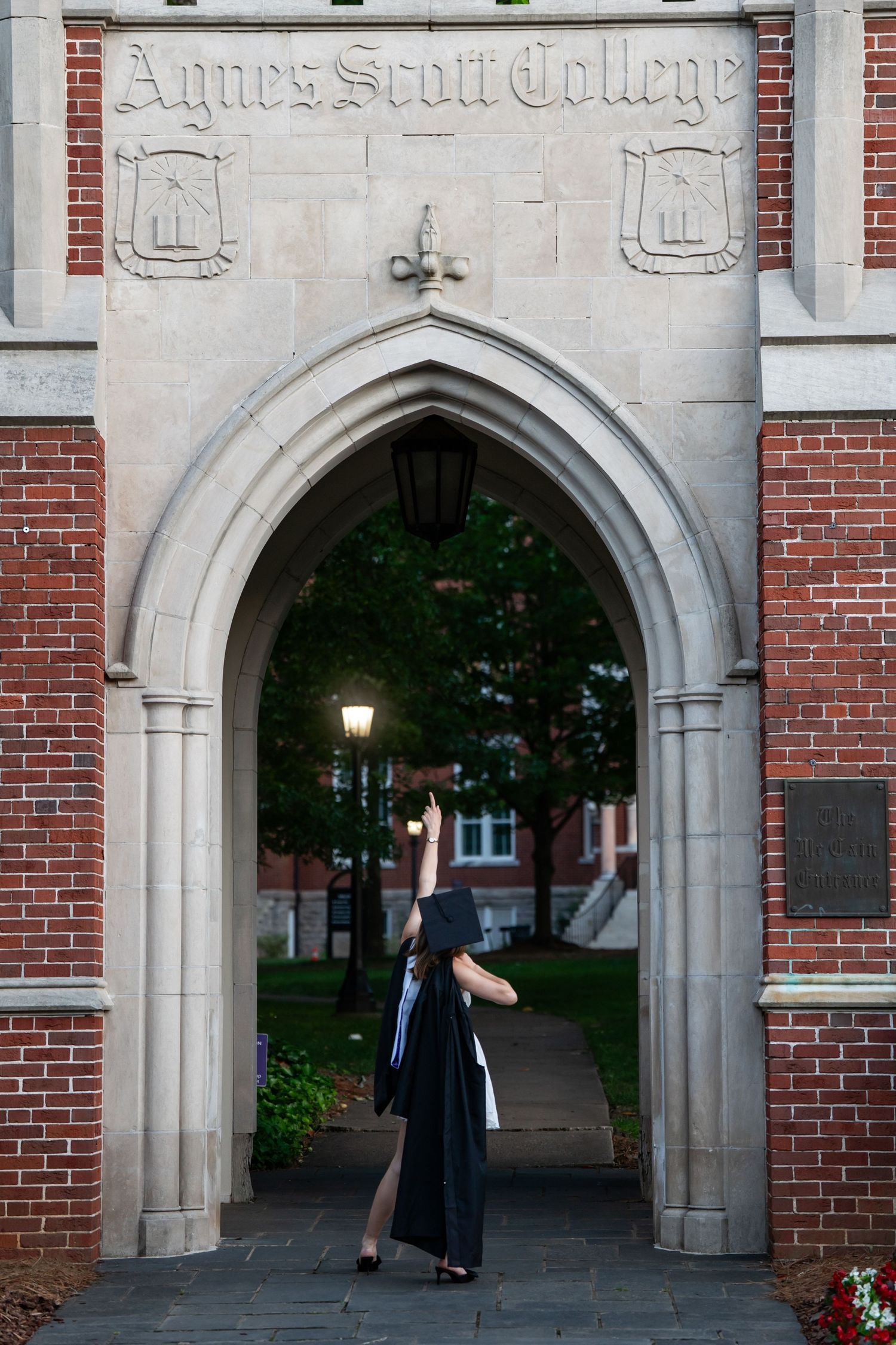 Graduate in black gown posing under Agnes Scott College stone archway at dusk.