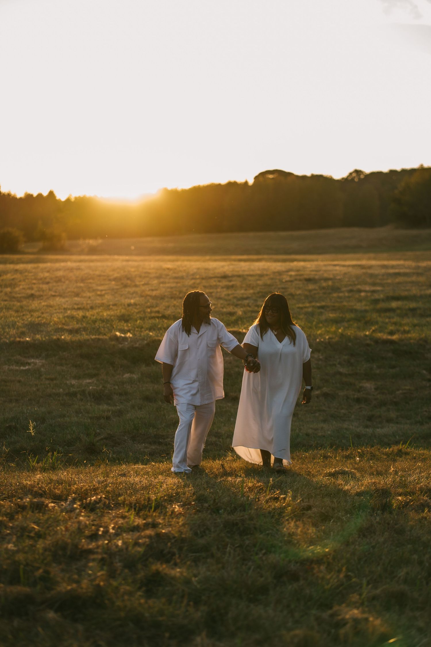 Couple dressed in white holding hands while walking through a sunlit meadow at golden hour.
