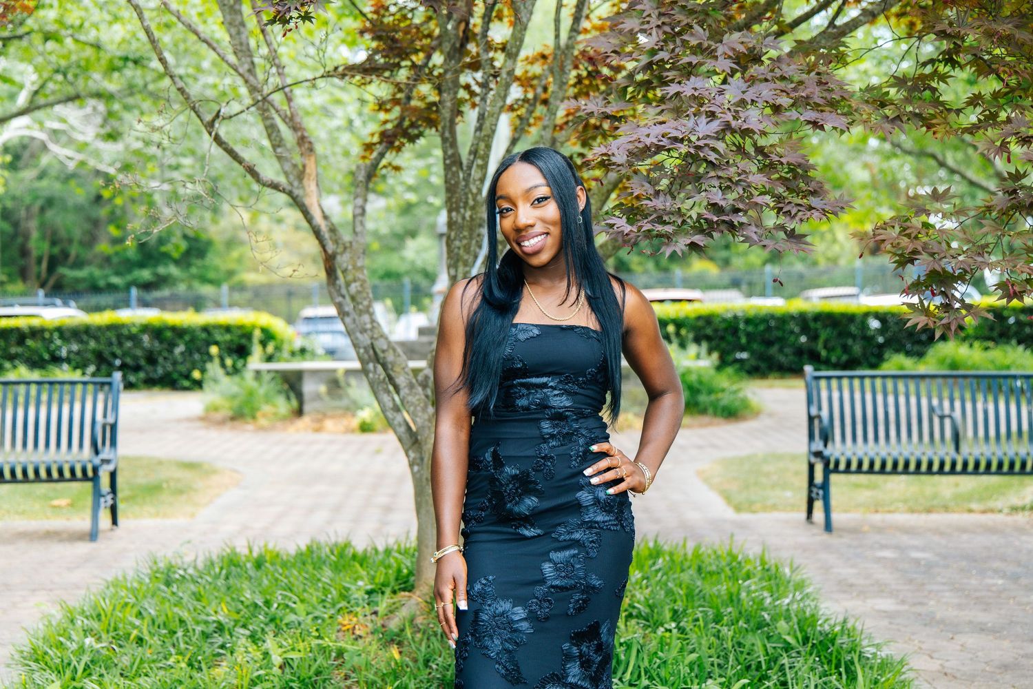Elegant woman in black fitted dress posing in lush garden park with colorful trees.