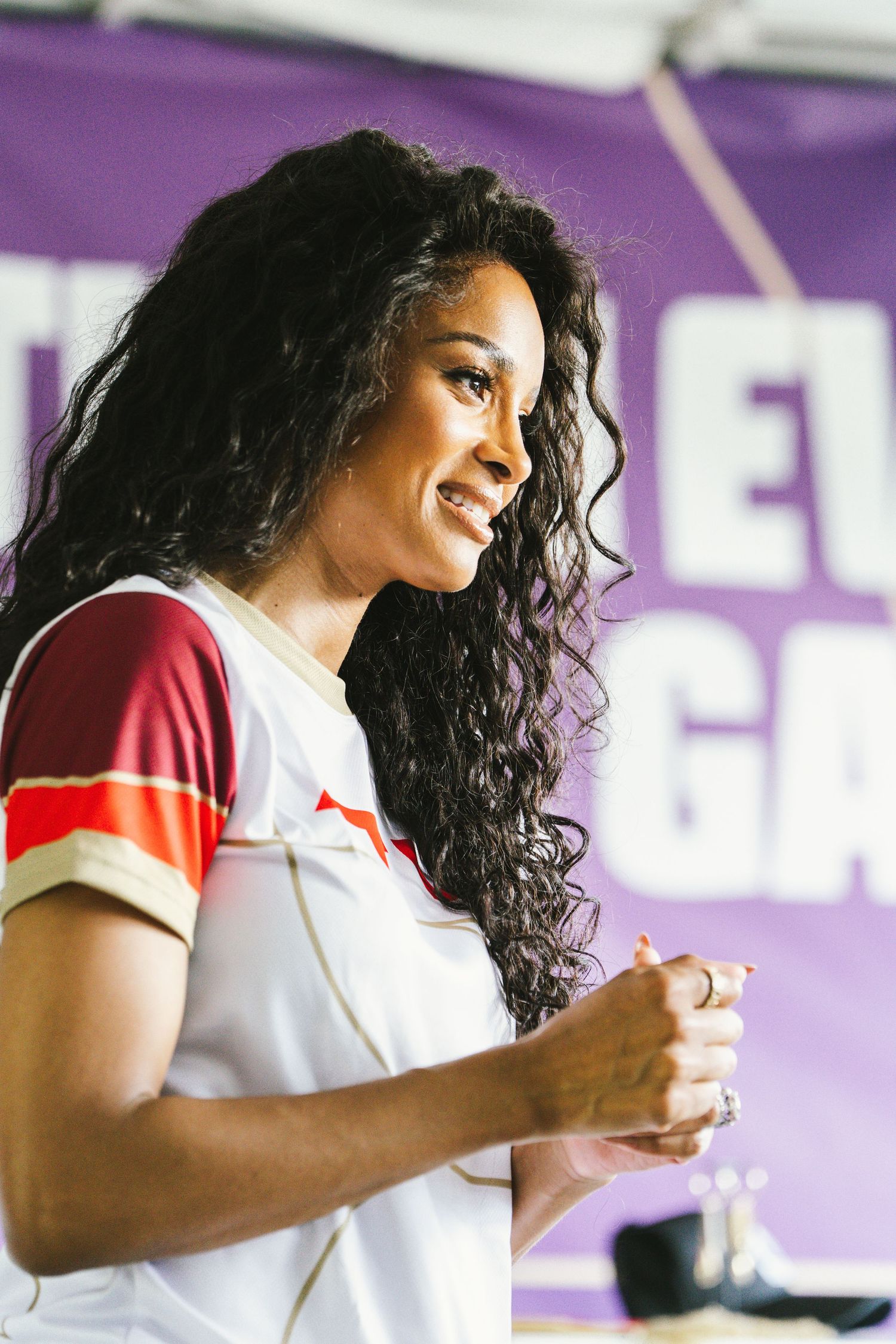 A smiling woman with curly hair speaking at an outdoor event with a purple banner.