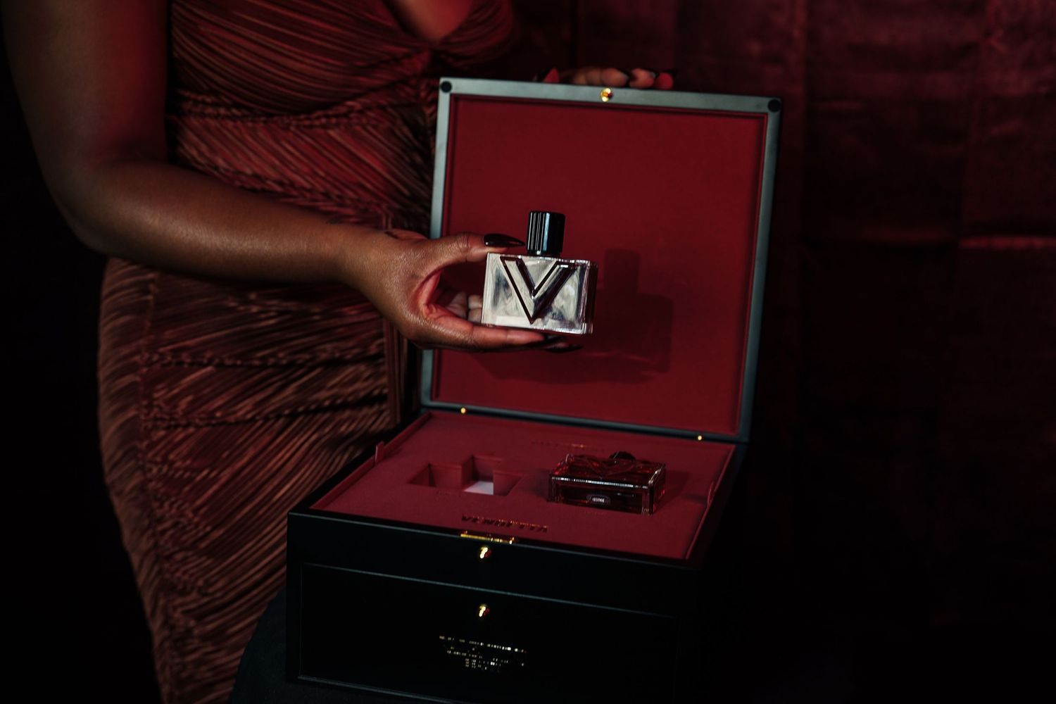 Woman in a red dress holds a luxury perfume bottle above an open black gift box with red velvet lining.
