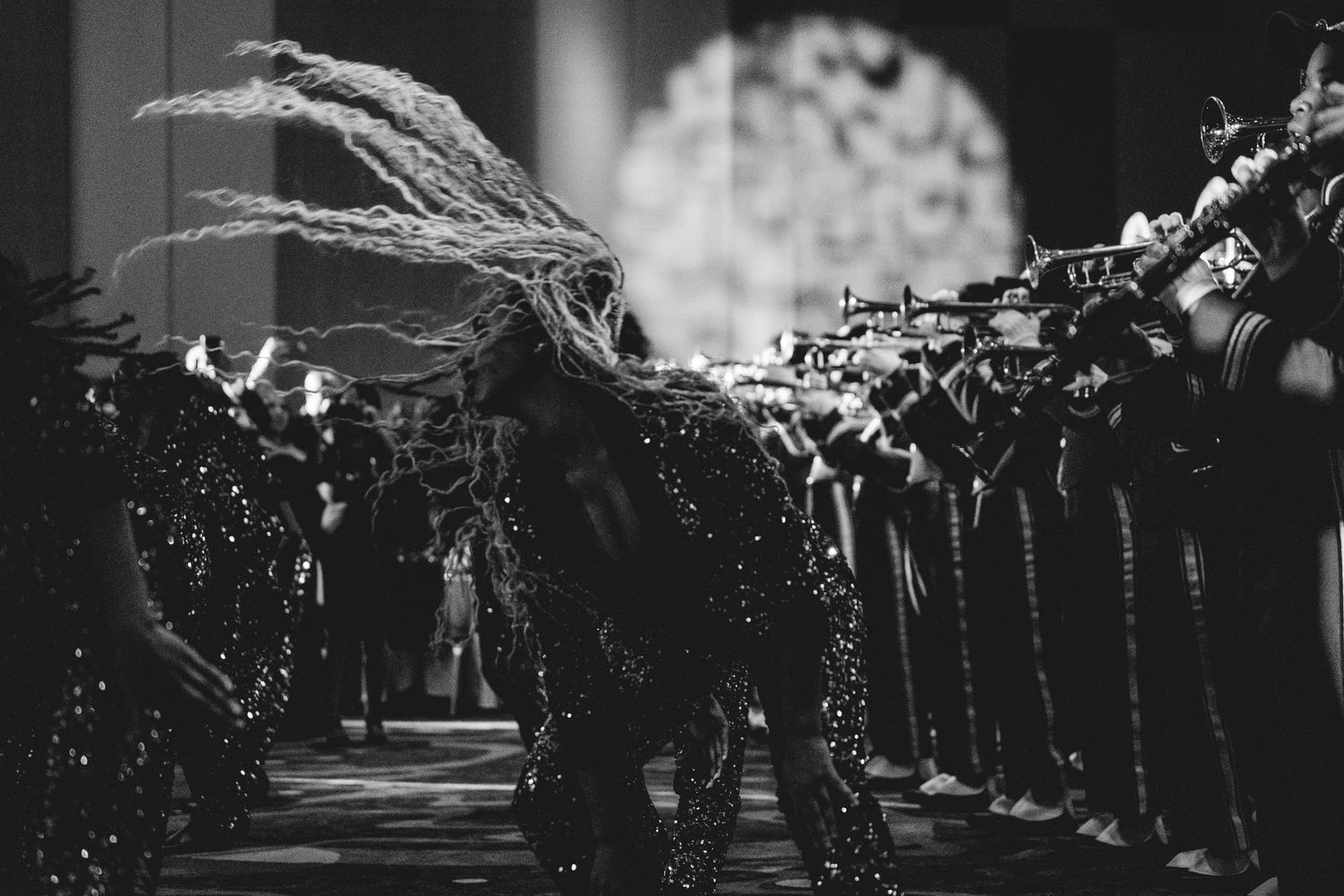 Black and white photo of feathered performer bowing before a line of brass musicians in sequined uniforms.