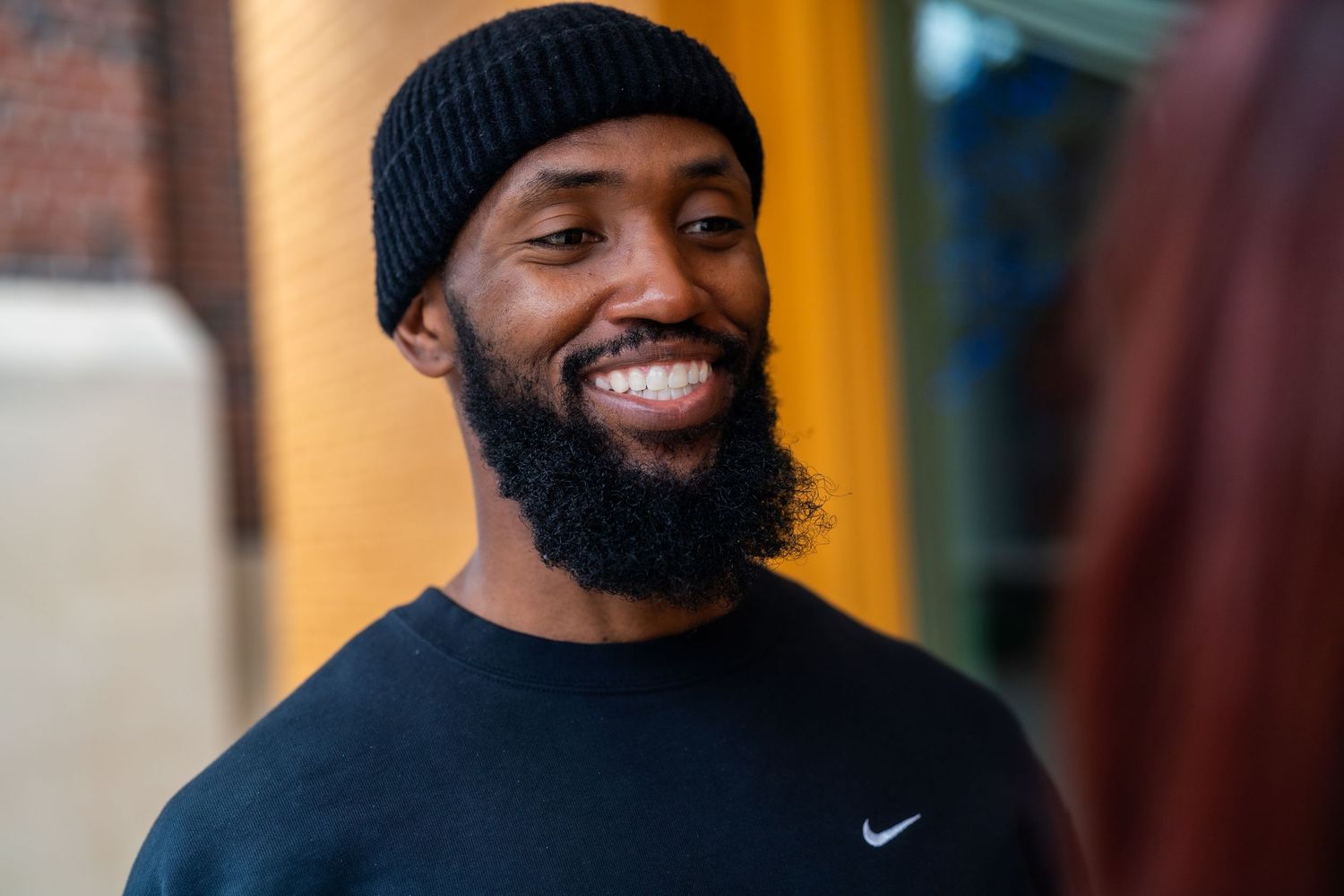 Smiling Black man with beard wearing black Nike beanie and shirt outdoors.