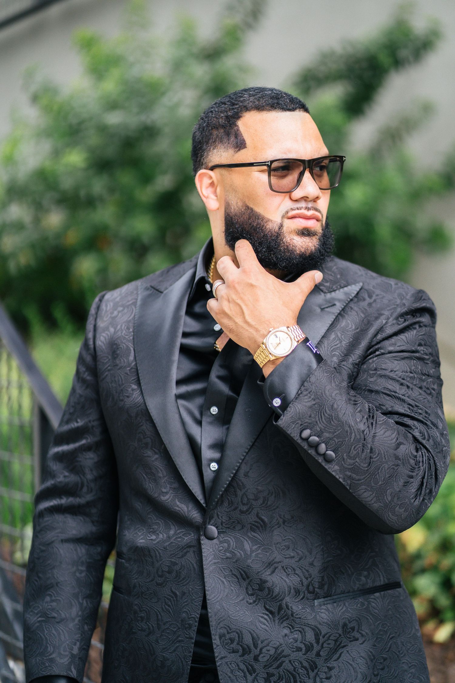 Confident bearded man in black patterned tuxedo jacket with gold watch stroking beard outdoors.