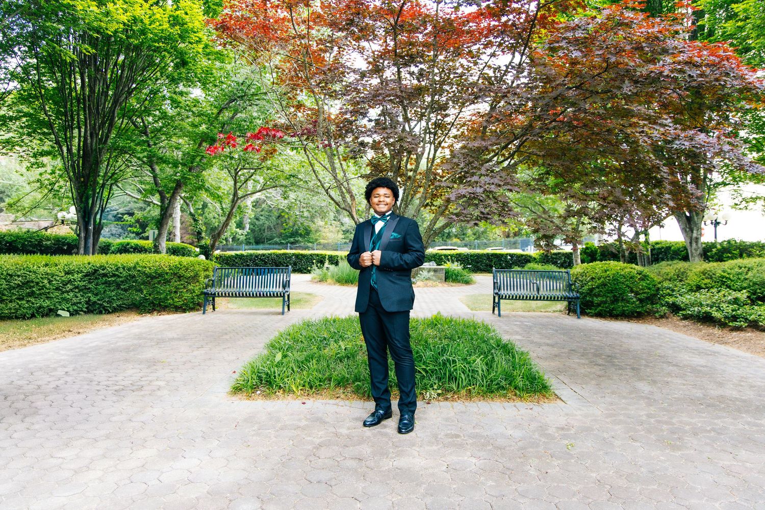 Man in black tuxedo with bow tie standing confidently in scenic park setting.