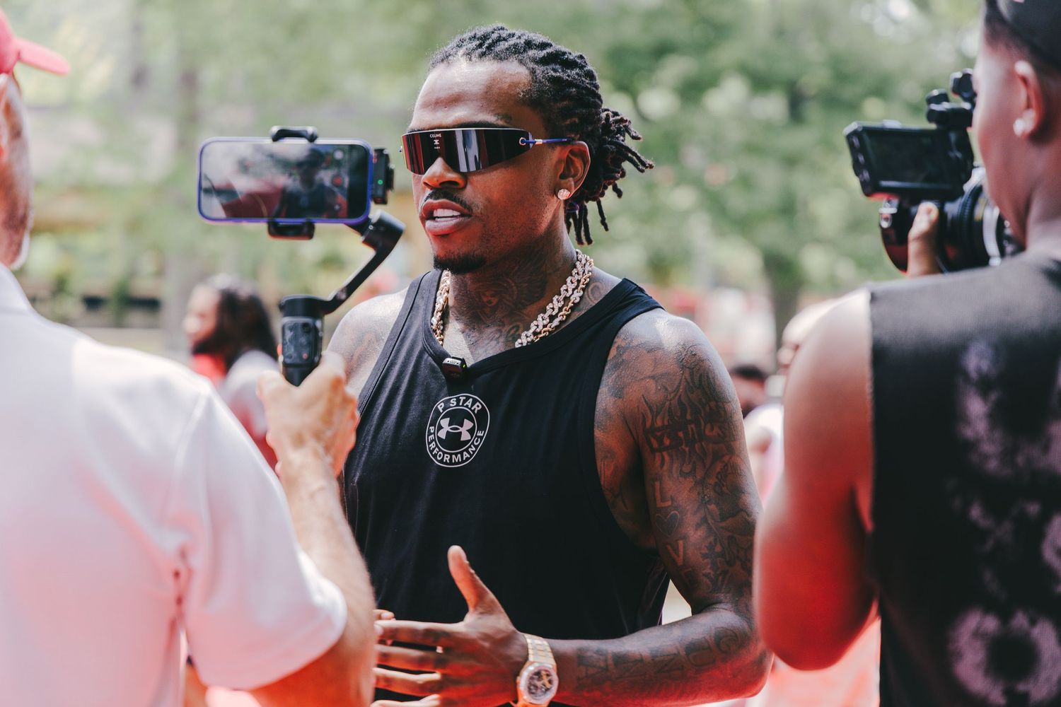Tattooed man in black sleeveless shirt and sunglasses with gold chain at outdoor sports event.