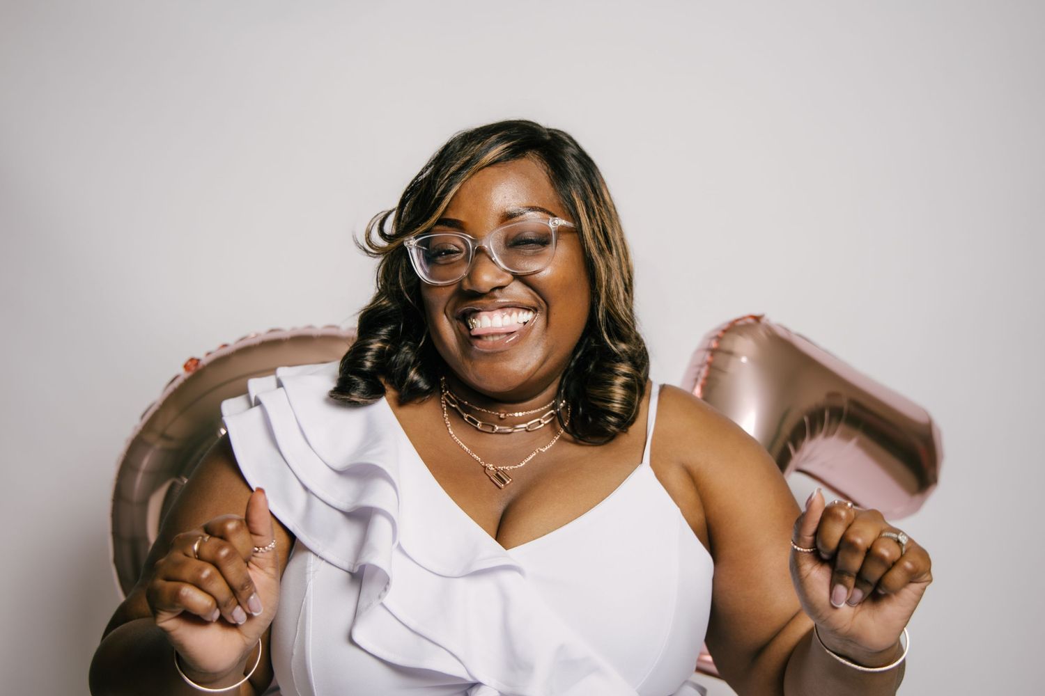 Joyful woman in white ruffled dress laughing with rose gold balloons in celebratory photo shoot.