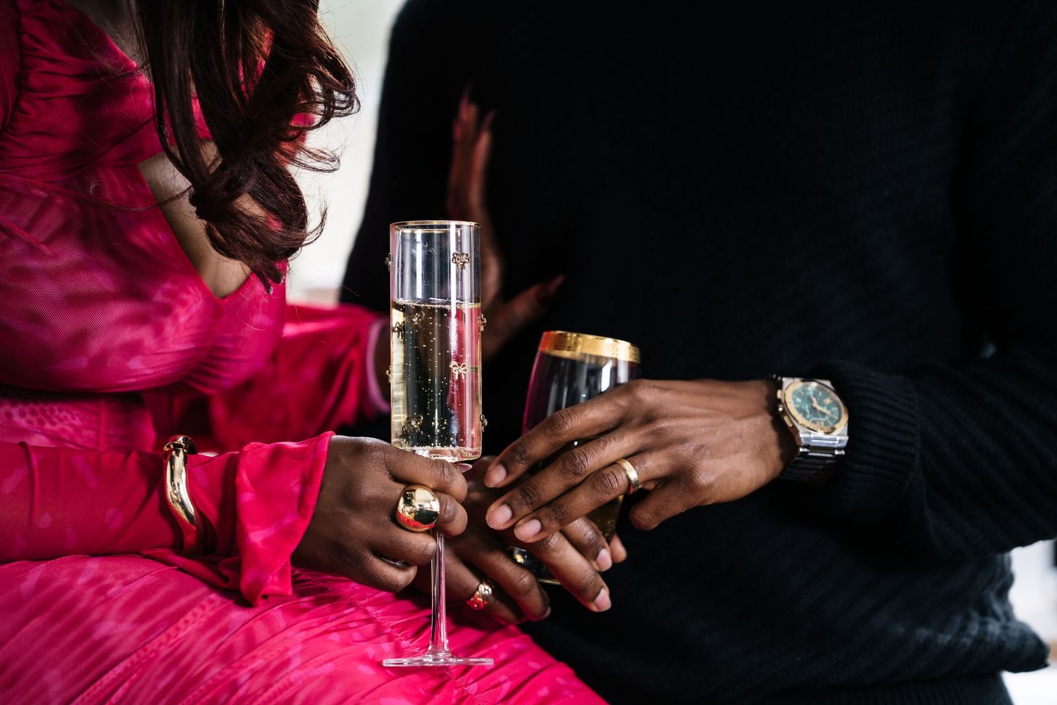Couple holding champagne flutes together, woman in pink dress and man in black suit with luxury watch.