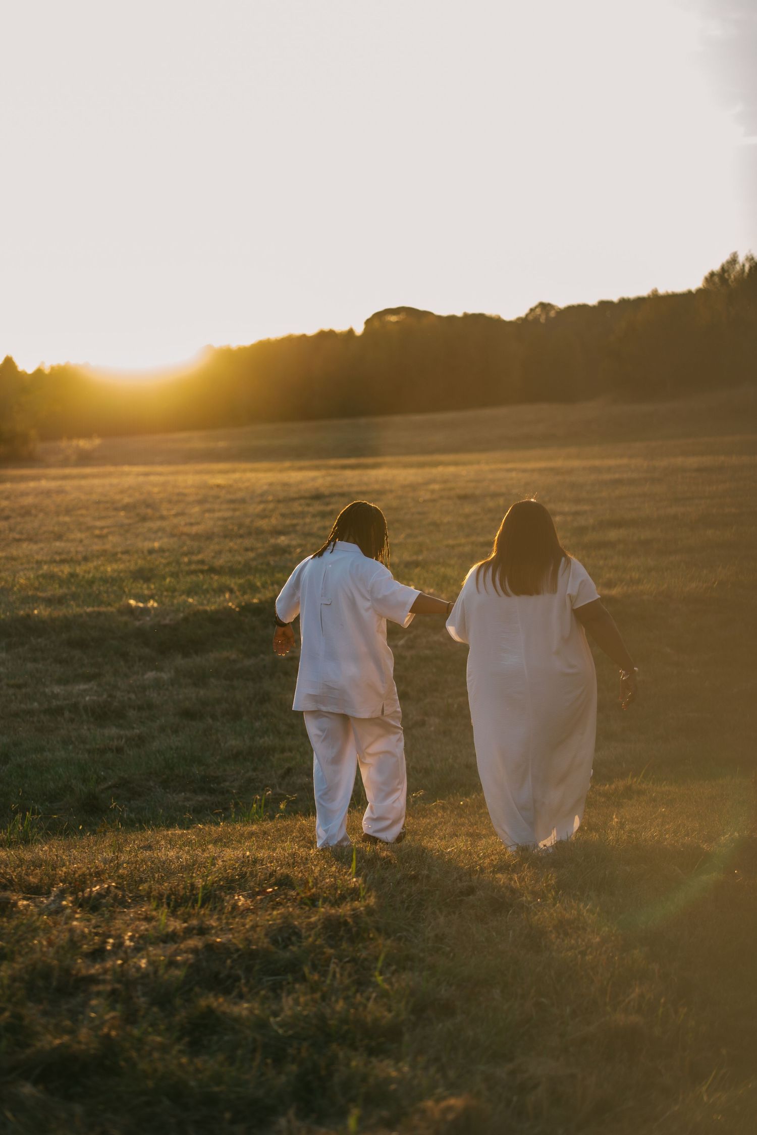 Two people in white outfits walking hand in hand through a golden field at sunset.
