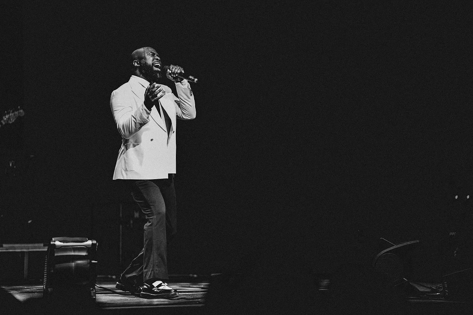 Black and white photo of a male singer performing on stage in a white suit jacket, holding a microphone.