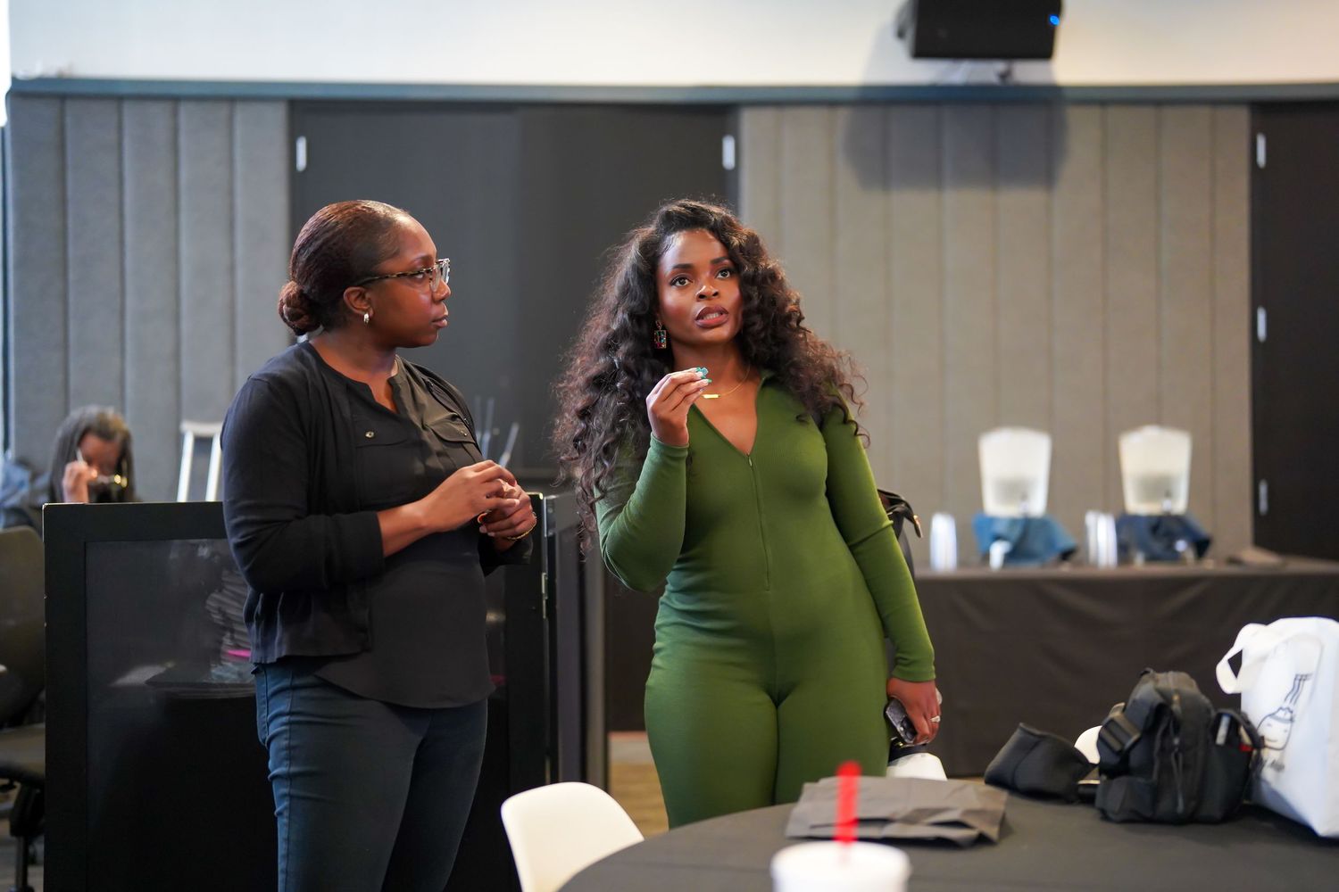 Two women engaged in conversation at an indoor event, one wearing a green jumpsuit.