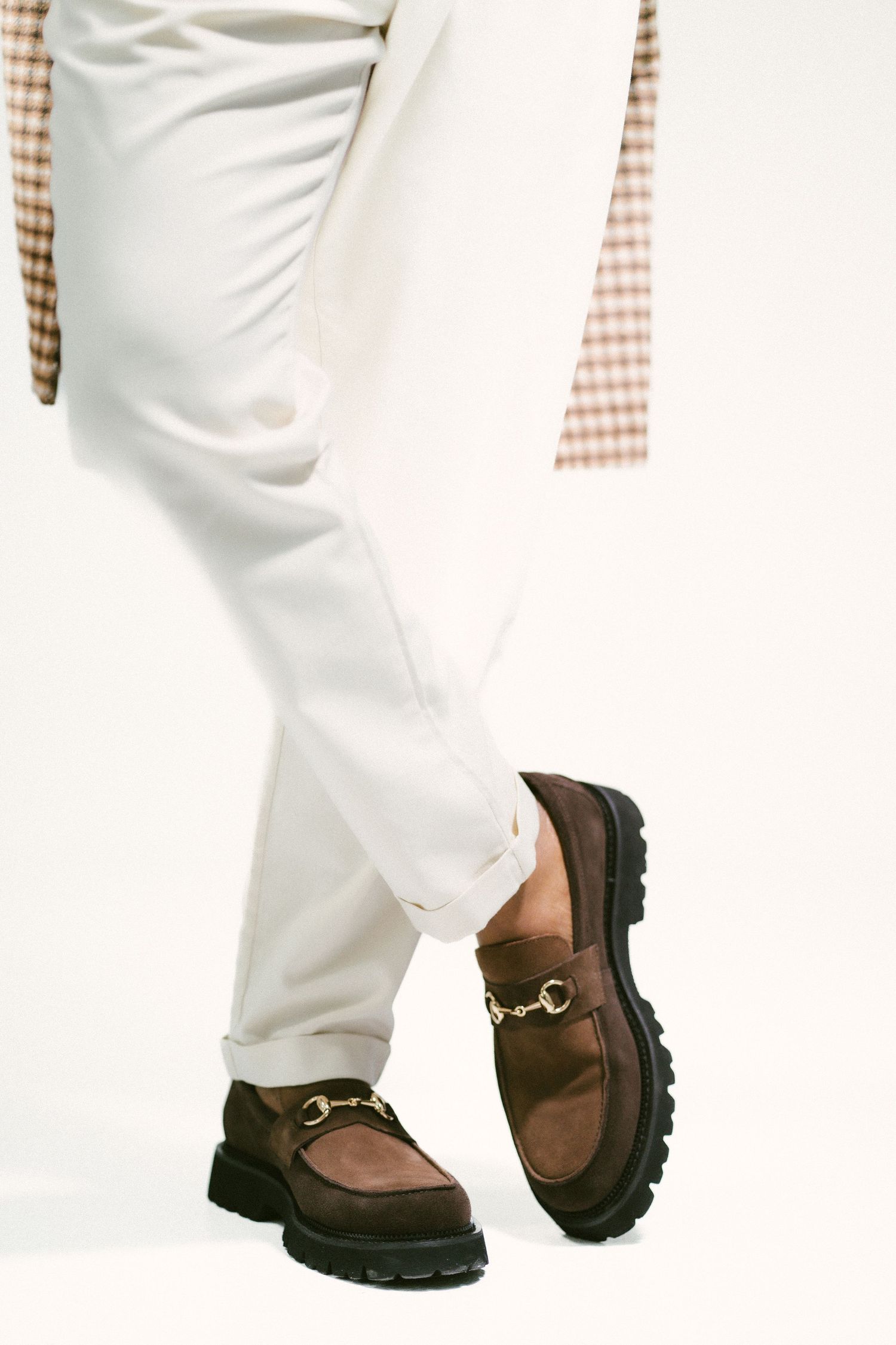 Brown suede horsebit loafers with chunky lug soles paired with white cuffed trousers and a plaid blazer.