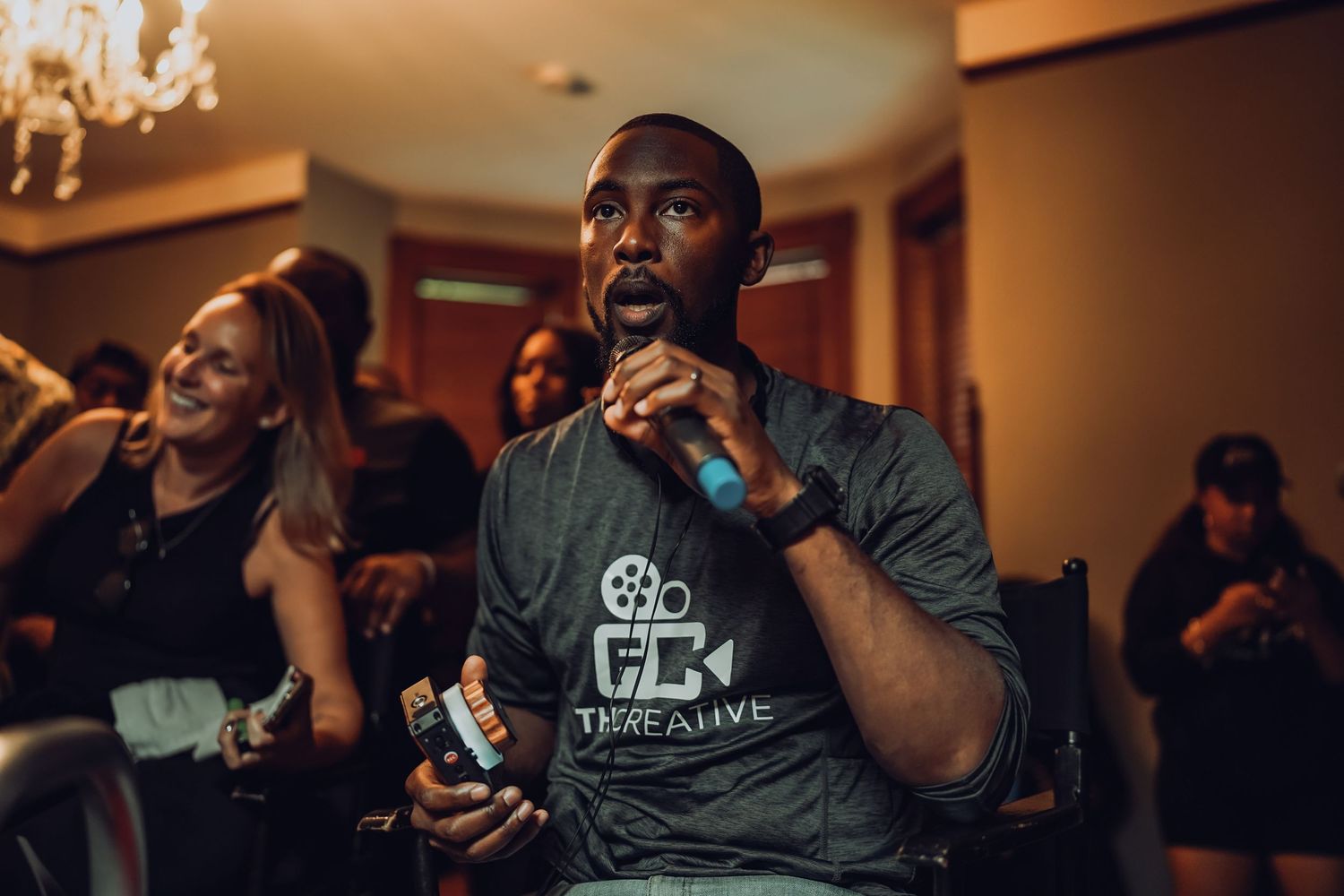 A man holding a microphone speaks at an indoor event wearing a gray ThreAlive film crew t-shirt.