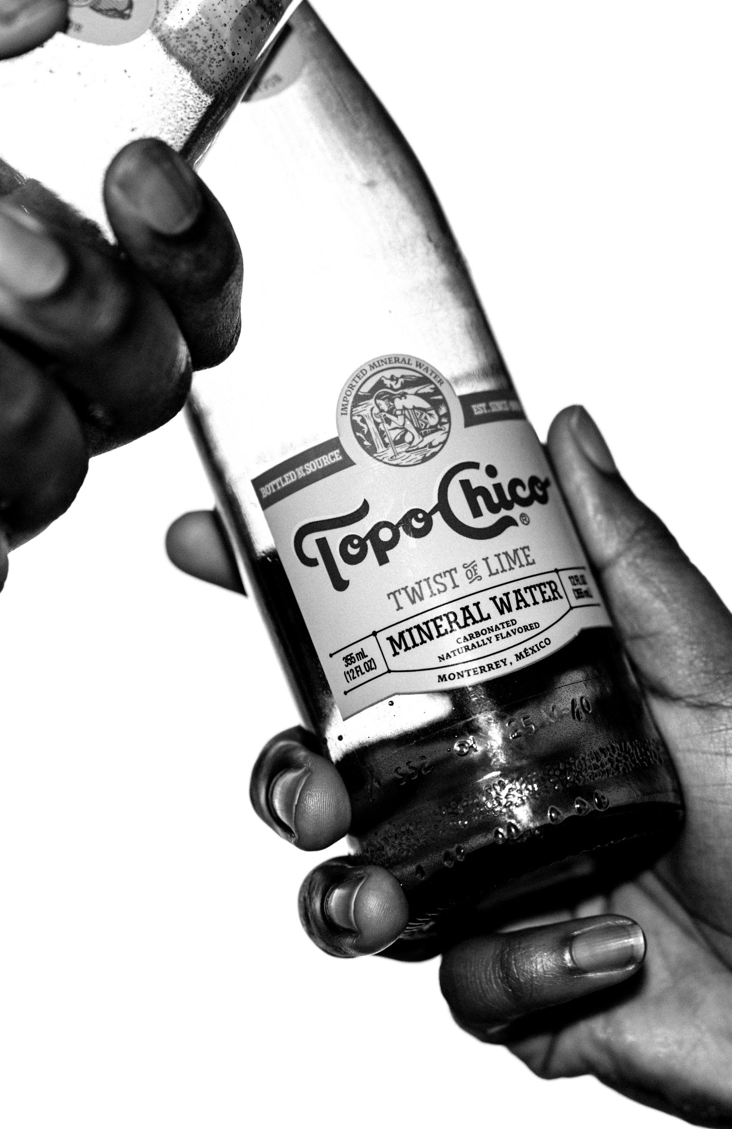 Black and white photo of two hands holding a Topo Chico Twist of Lime mineral water bottle.