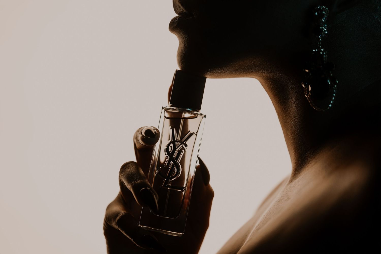 Silhouetted woman in profile holding a YSL perfume bottle to her lips, wearing a sparkling drop earring against a warm sepia background.