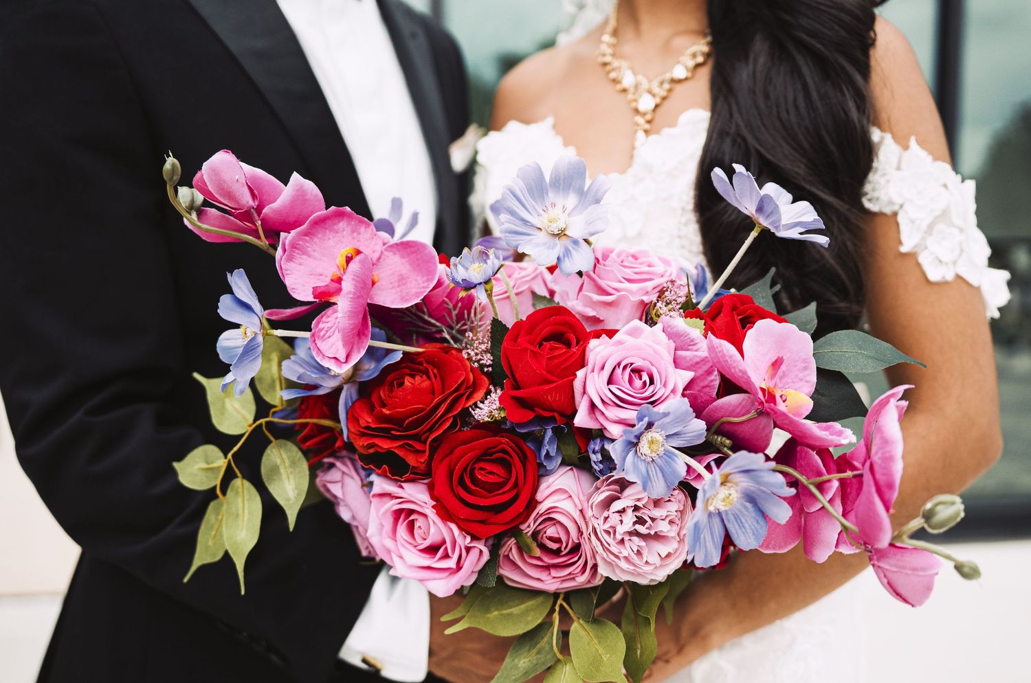 Bride and groom holding a colorful wedding bouquet with red roses and pink orchids outdoors.
