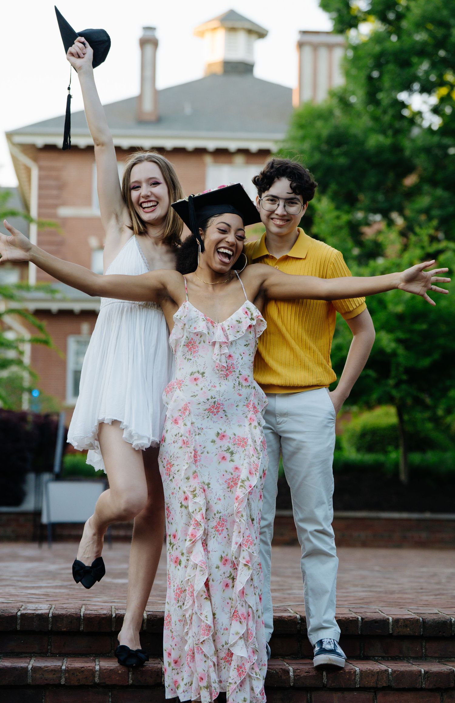 Three joyful graduates celebrate outdoors near a brick building, with one wearing a cap and gown.