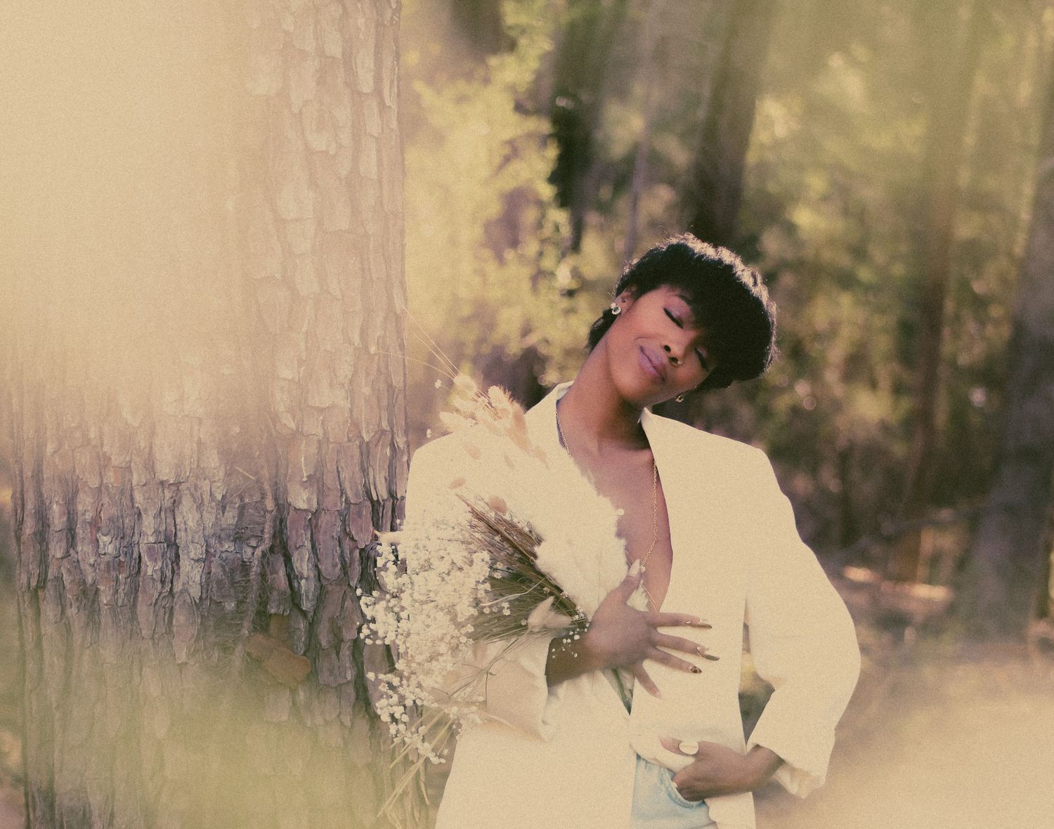 A woman in a white blazer holds white flowers while leaning against a tree in a softly lit forest setting.