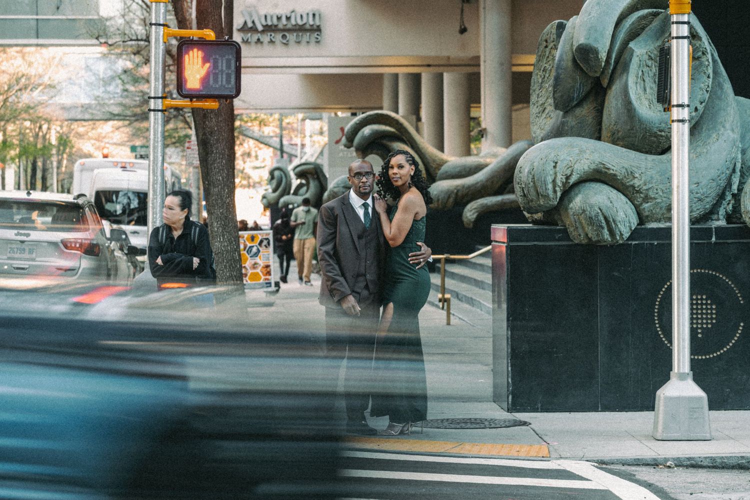 Couple posing near a large sculpture on a busy city street with traffic passing by.