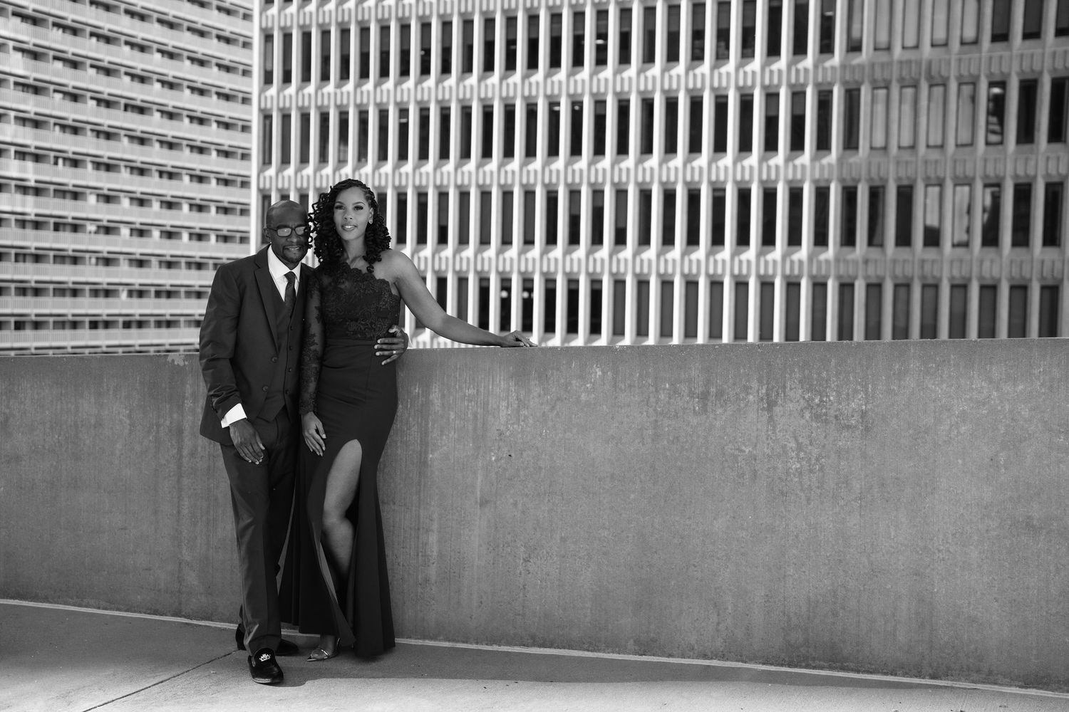 Elegant couple in formal attire posing against urban building wall in black and white photo.